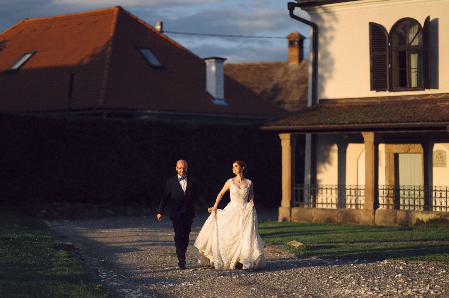 Wedding couple walks across a grassy field at sunset near rural buildings as their gowns flow in the breeze.