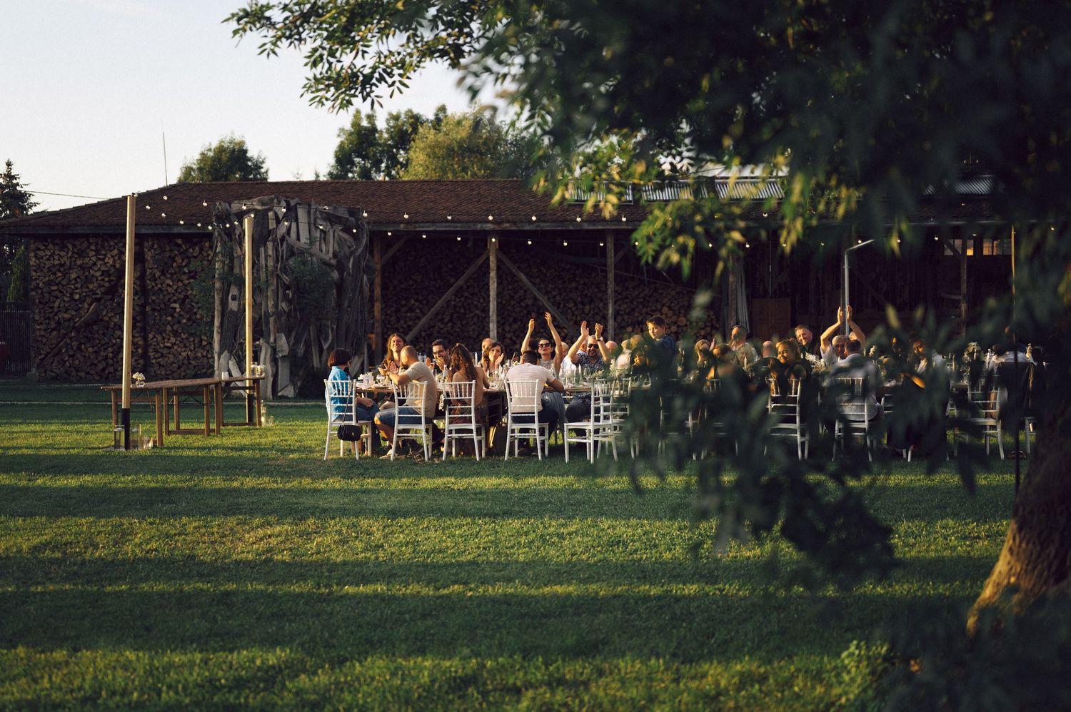Guests dine at long tables under rustic wooden pergolas during an outdoor garden reception at sunset.