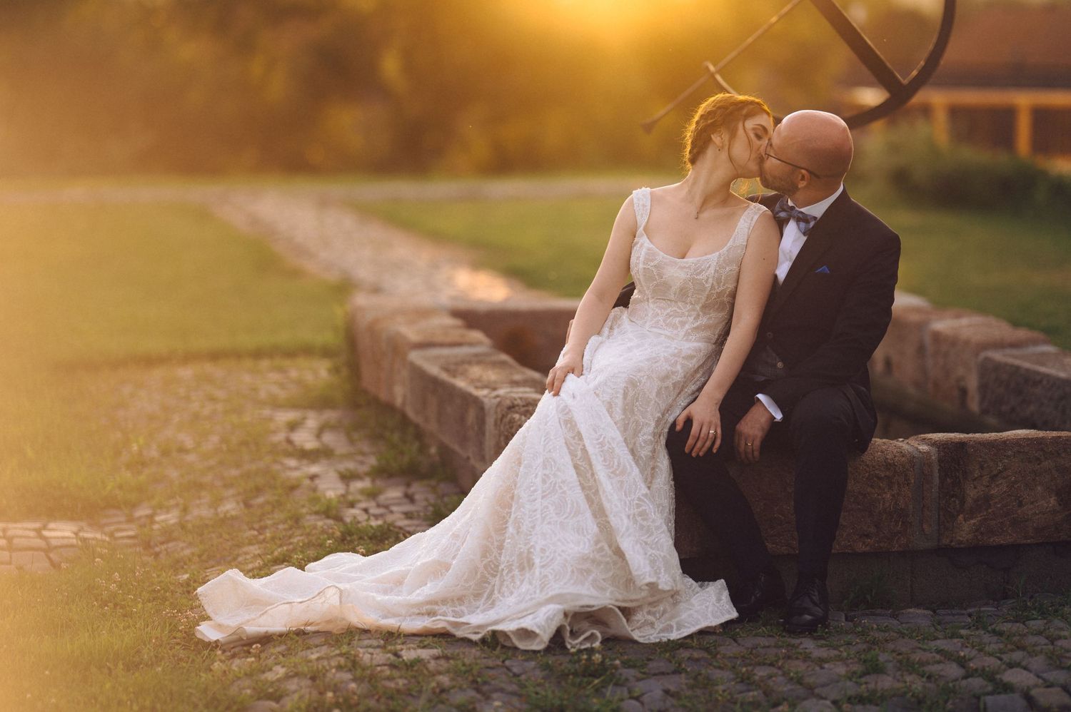 Newlywed couple sits on stone wall during golden hour sunset in romantic wedding portrait.