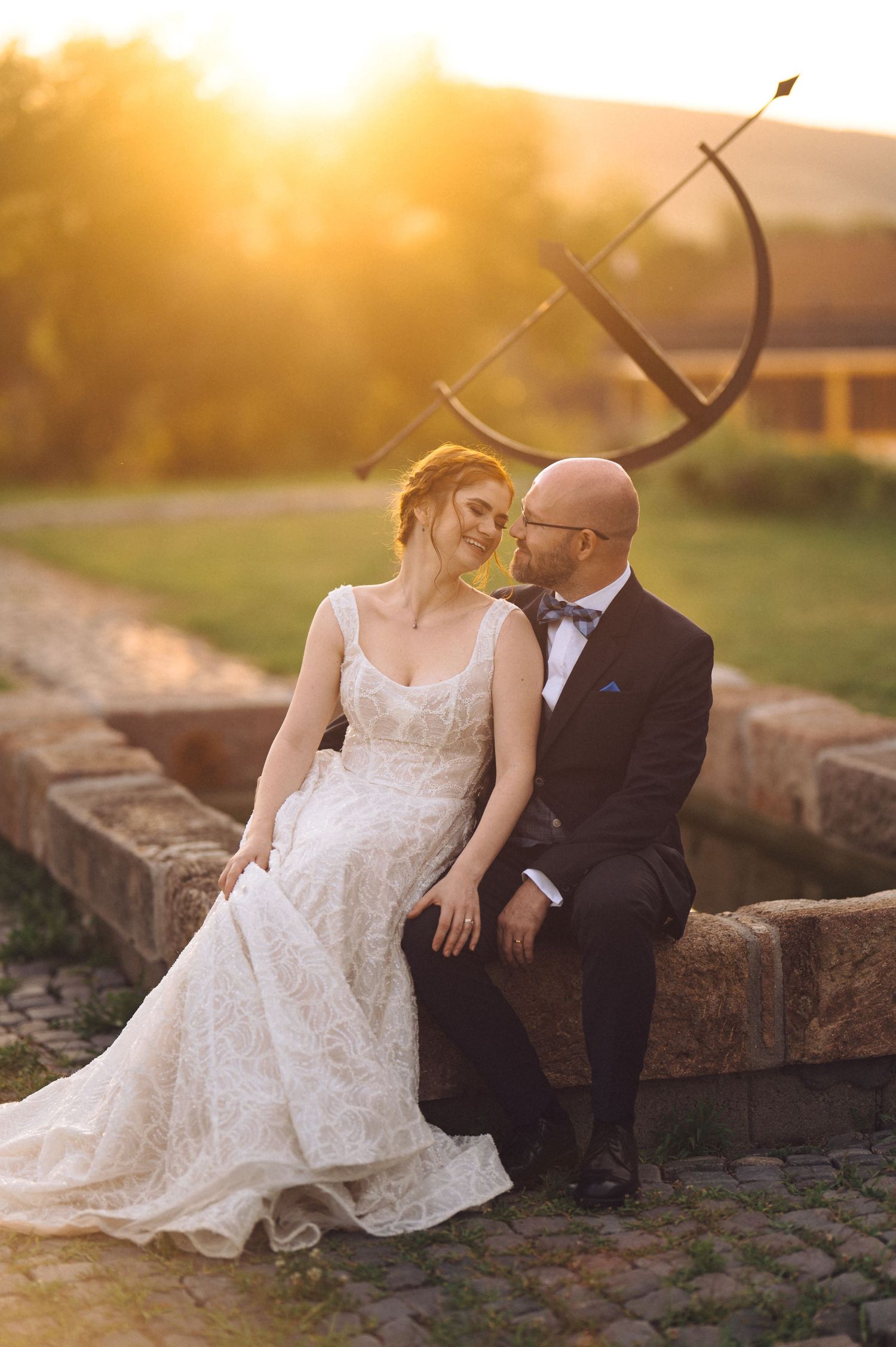 A couple sits together at sunset with a beautiful lace wedding dress and classic black tuxedo.
