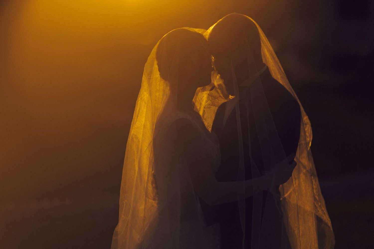 Silhouetted wedding couple embraces under veil with warm orange sunset lighting.