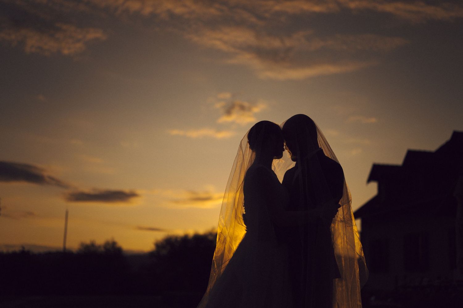 Dramatic sunset silhouette of wedding couple sharing intimate moment under flowing veil.