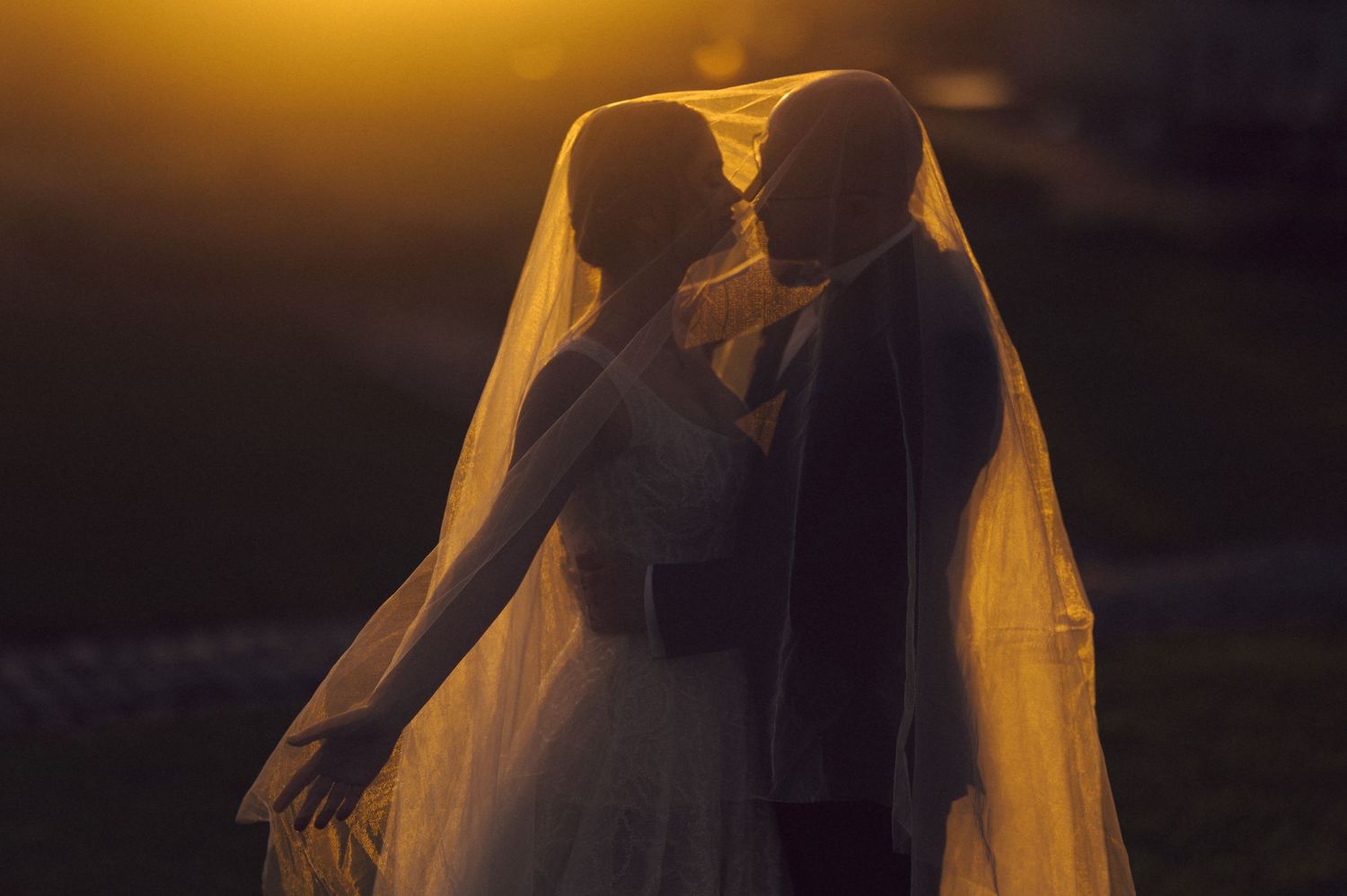A silhouetted bride and groom share an intimate moment under flowing veil during golden hour.