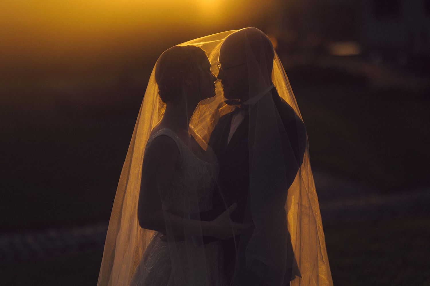 Romantic silhouette of bride and groom beneath flowing veil at sunset.