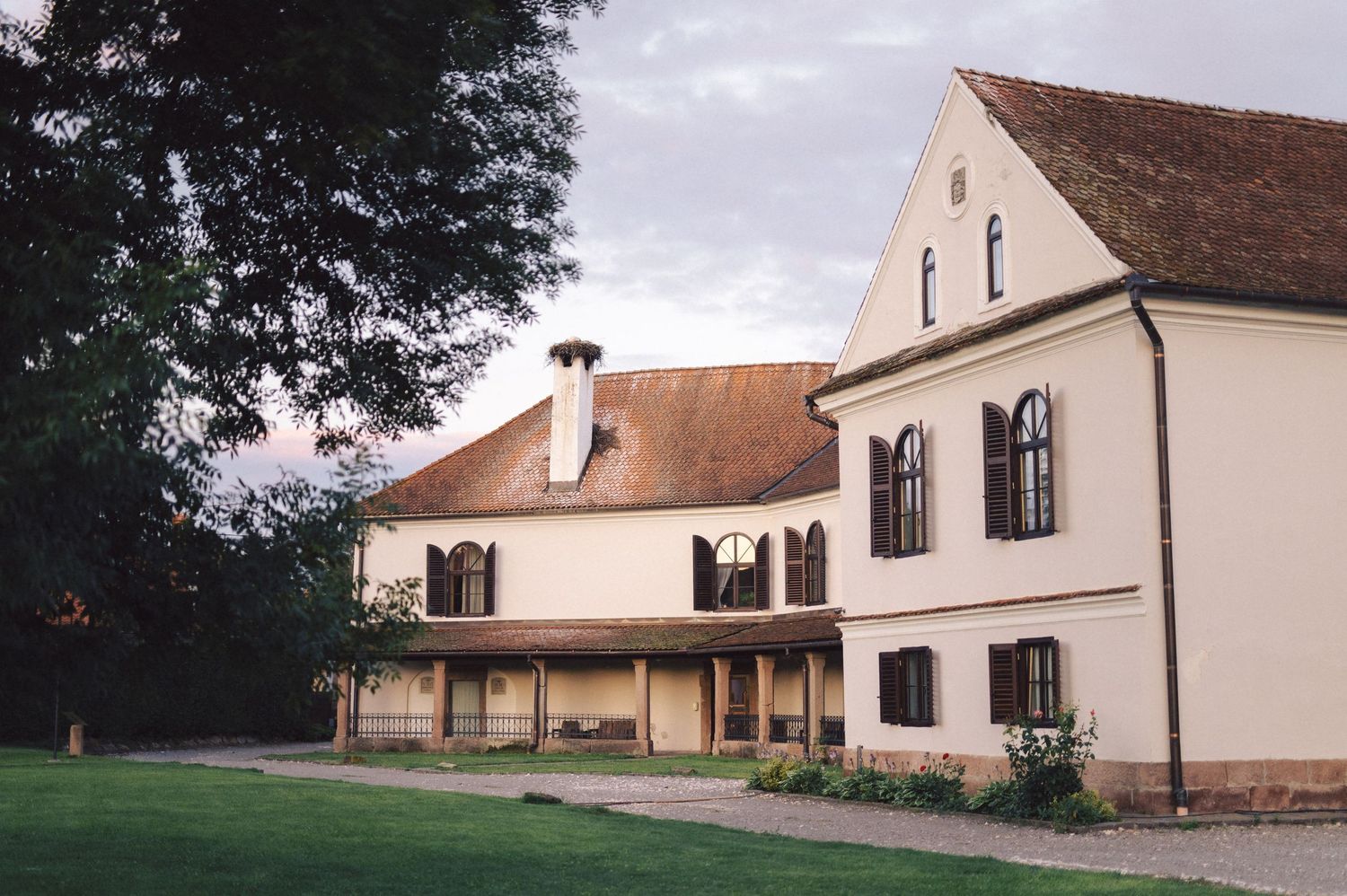 A traditional cream-colored manor house with brown roof and multiple windows in a garden setting.