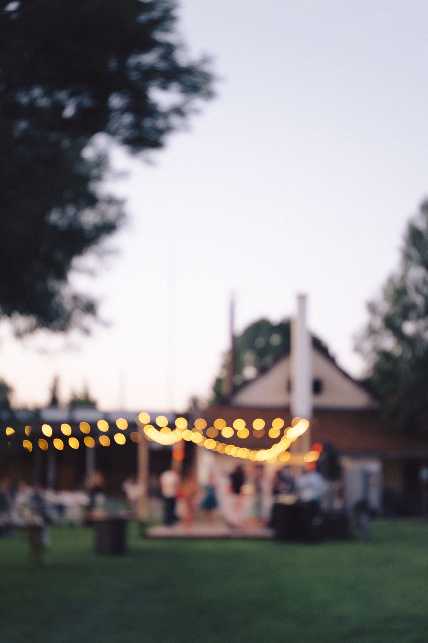 String lights illuminate an outdoor backyard party setting at dusk with a house silhouette in the background.