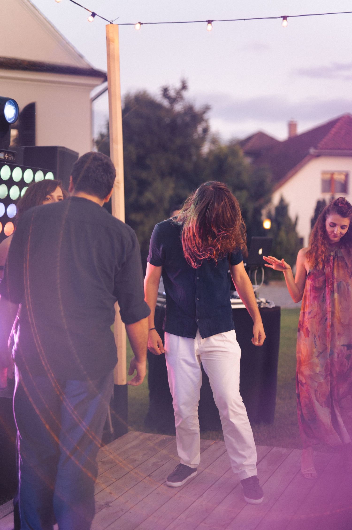People dancing at an outdoor party beneath string lights with a purple-tinted ambiance in the evening.