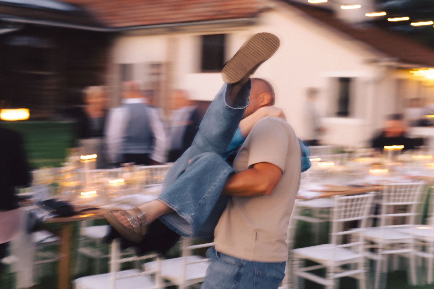 Blurred motion shot of people dancing together at an outdoor wedding reception with white chairs and tables.