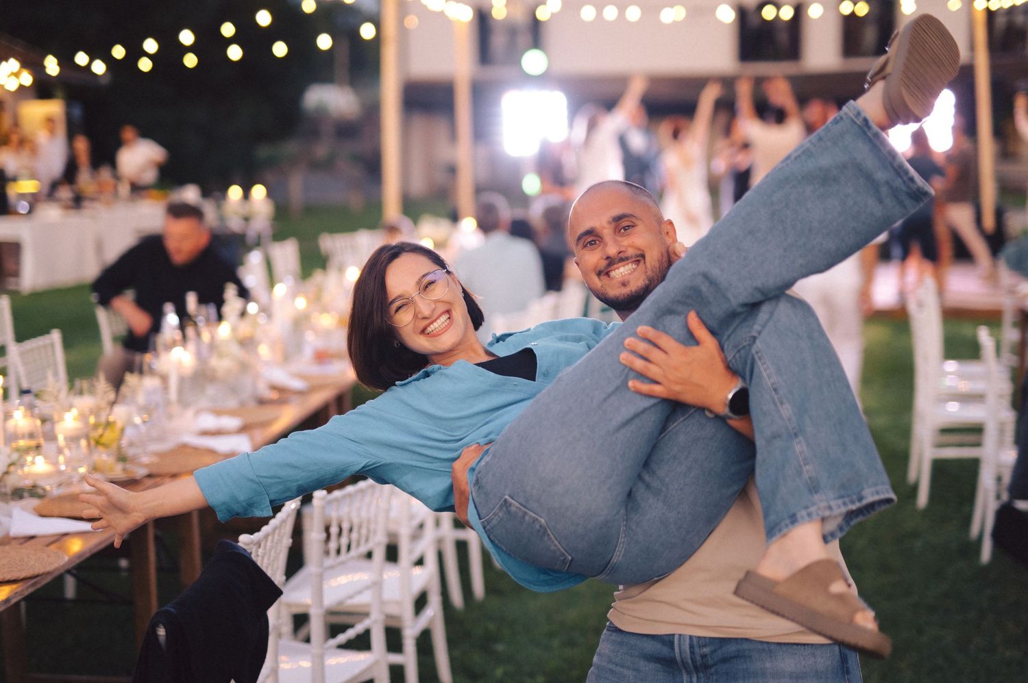 String lights illuminate a lively outdoor wedding reception as guests dance and celebrate on a warm evening.