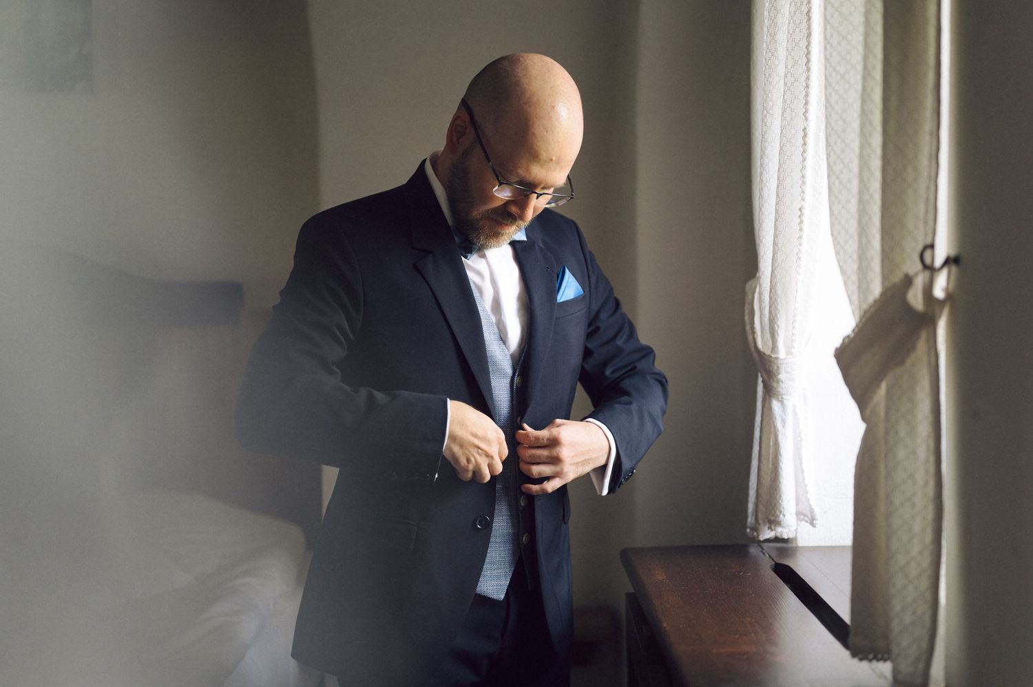 A person in a navy suit adjusts their buttons while standing near a sunlit window.