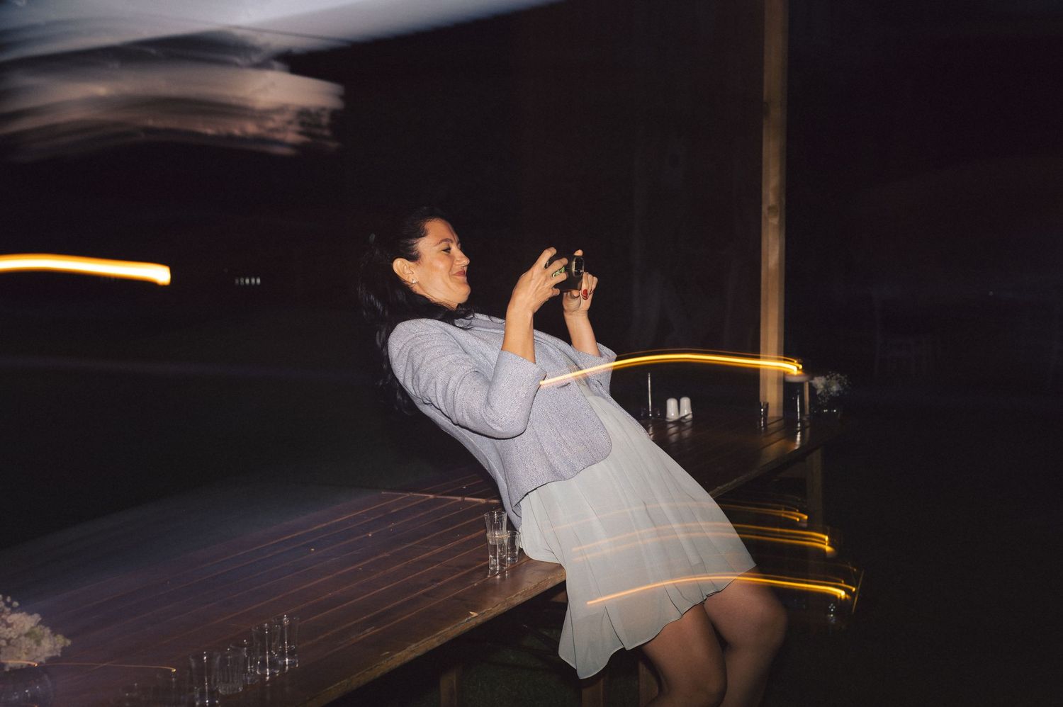 A person in a grey dress takes a photograph at night, with light trails creating a dramatic streaked effect.