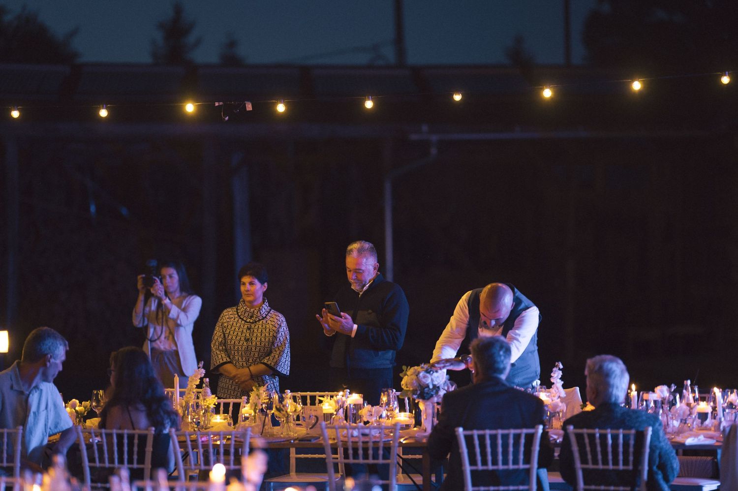 People gather around an outdoor dining table at night with string lights illuminating the evening atmosphere.