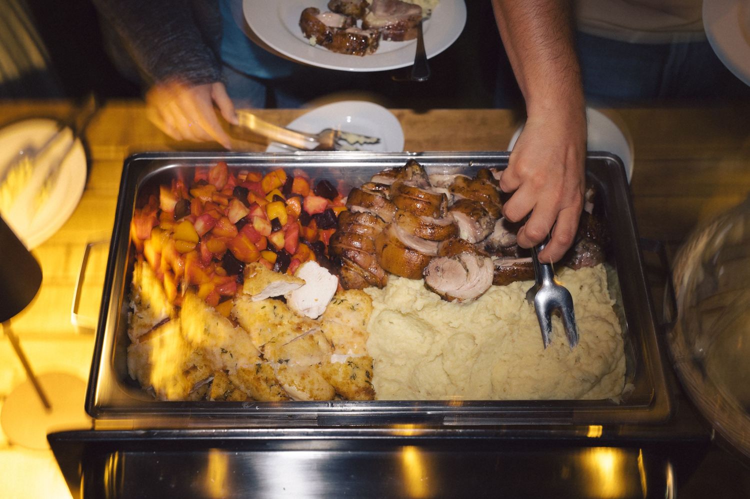 Food cooking in metal serving trays at a buffet or catering event with hands reaching to serve portions.