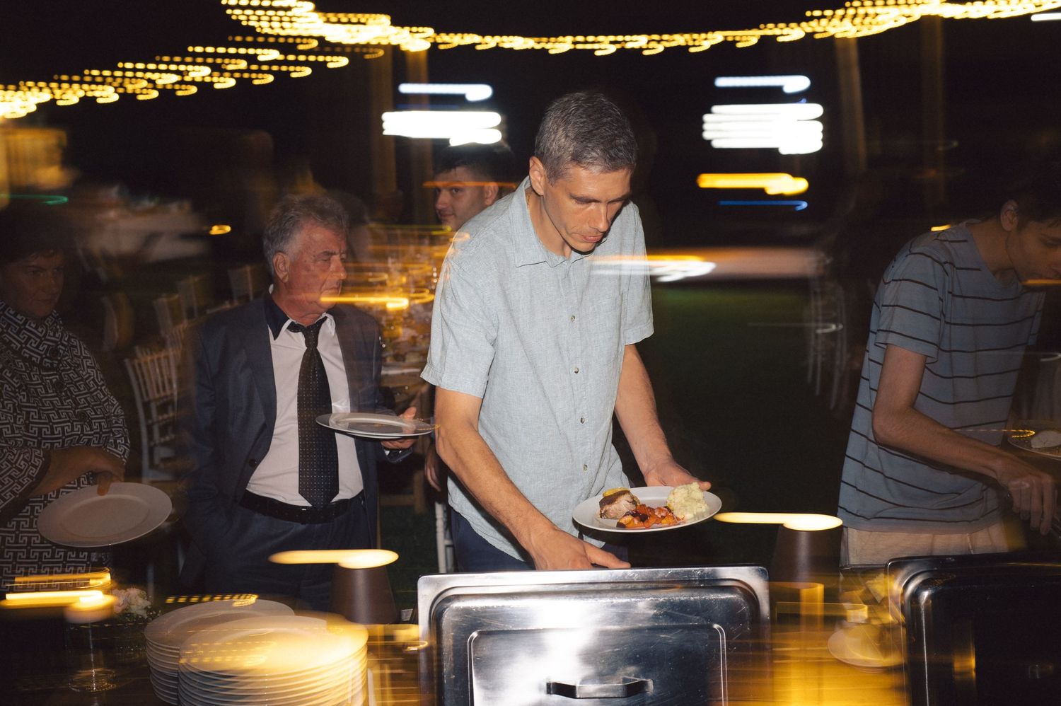 People gather around a buffet table with warm lighting and decorative string lights overhead during an evening event.