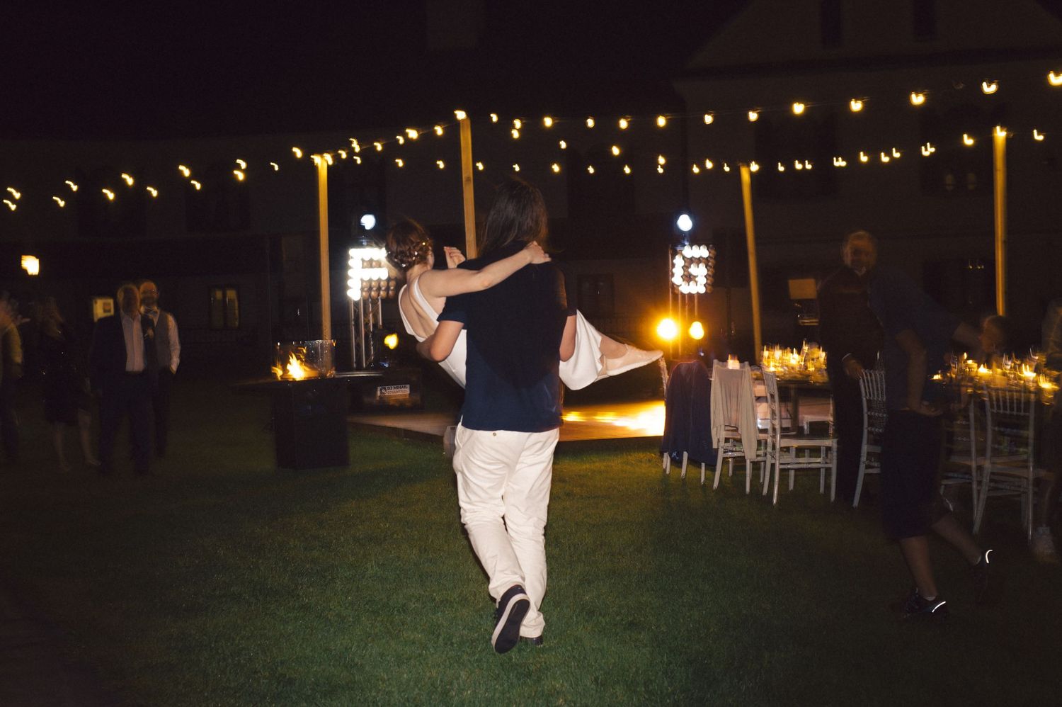 Couple dancing together at an outdoor evening reception with string lights overhead on a grassy lawn.