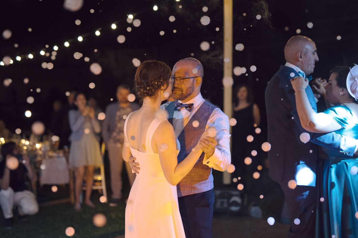 A couple dances outdoors at a nighttime wedding reception with sparkling lights illuminating the romantic atmosphere.