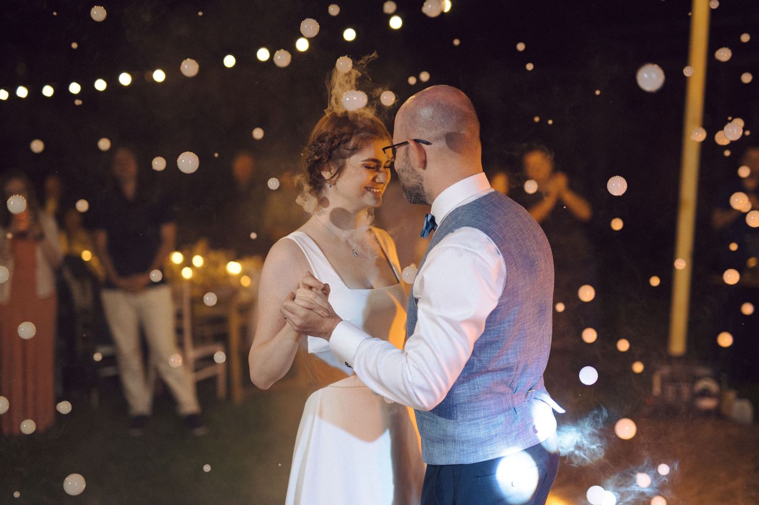 A couple shares their first dance as newlyweds under romantic string lights at an outdoor evening reception.