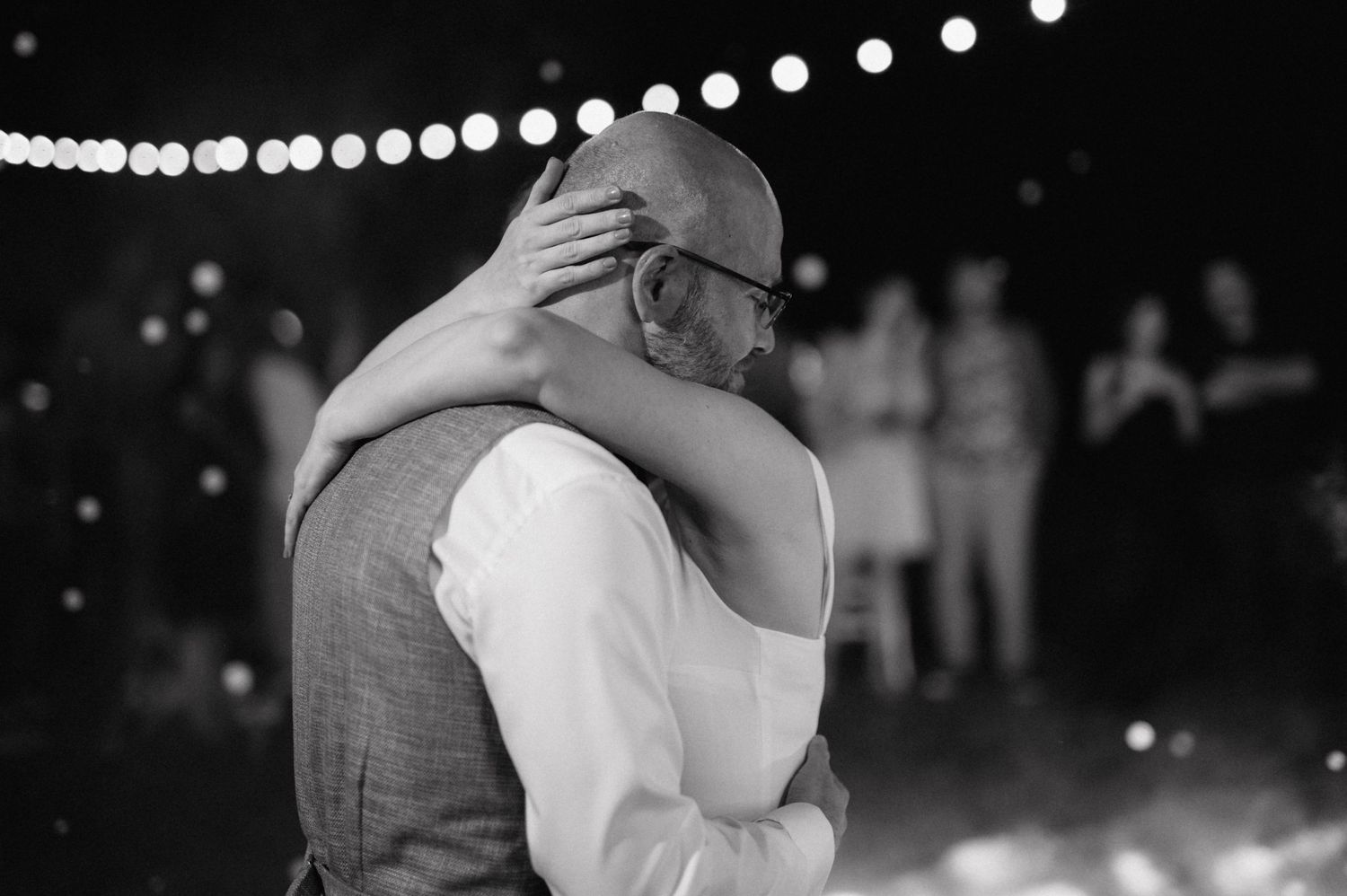 A tender moment is captured in black and white under string lights at an outdoor evening event.