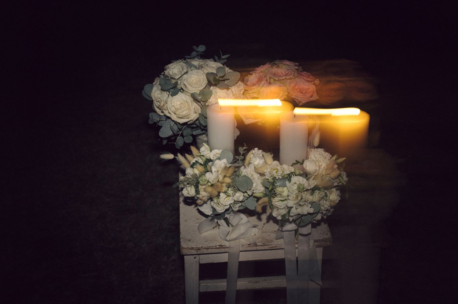 Lit white candles glow warmly beside white flowers arranged in a wooden box against a dark background.