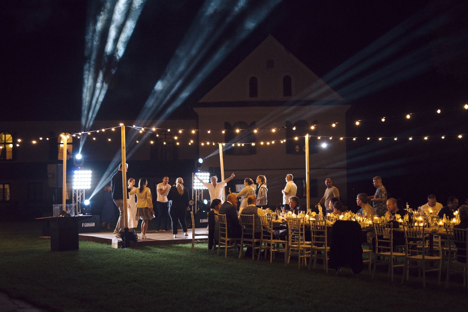 Elegant outdoor wedding reception under string lights at night with dramatic beams of light illuminating the scene.