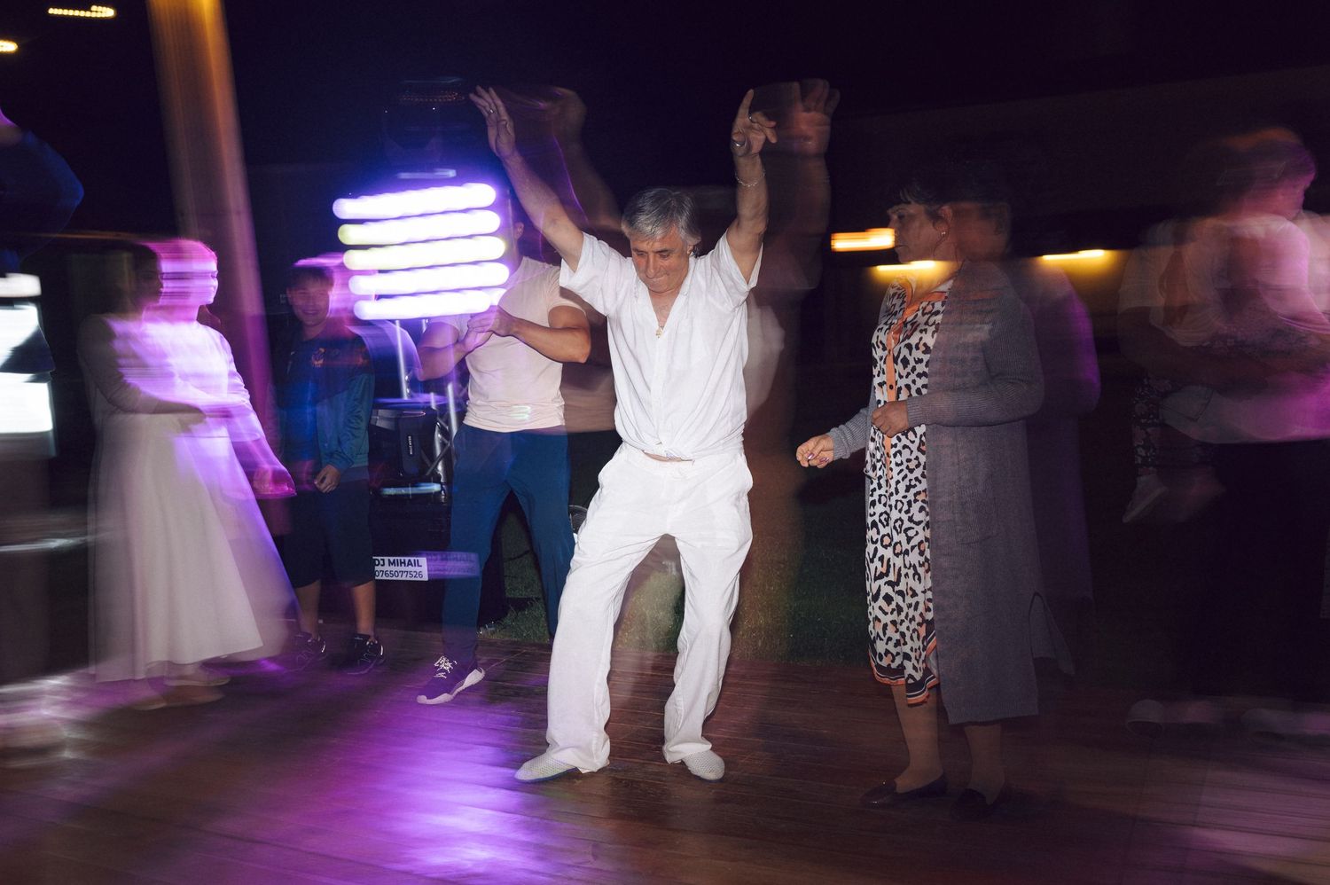 Dancer in all white outfit dancing energetically on a dimly lit dance floor with purple lighting and other guests around.