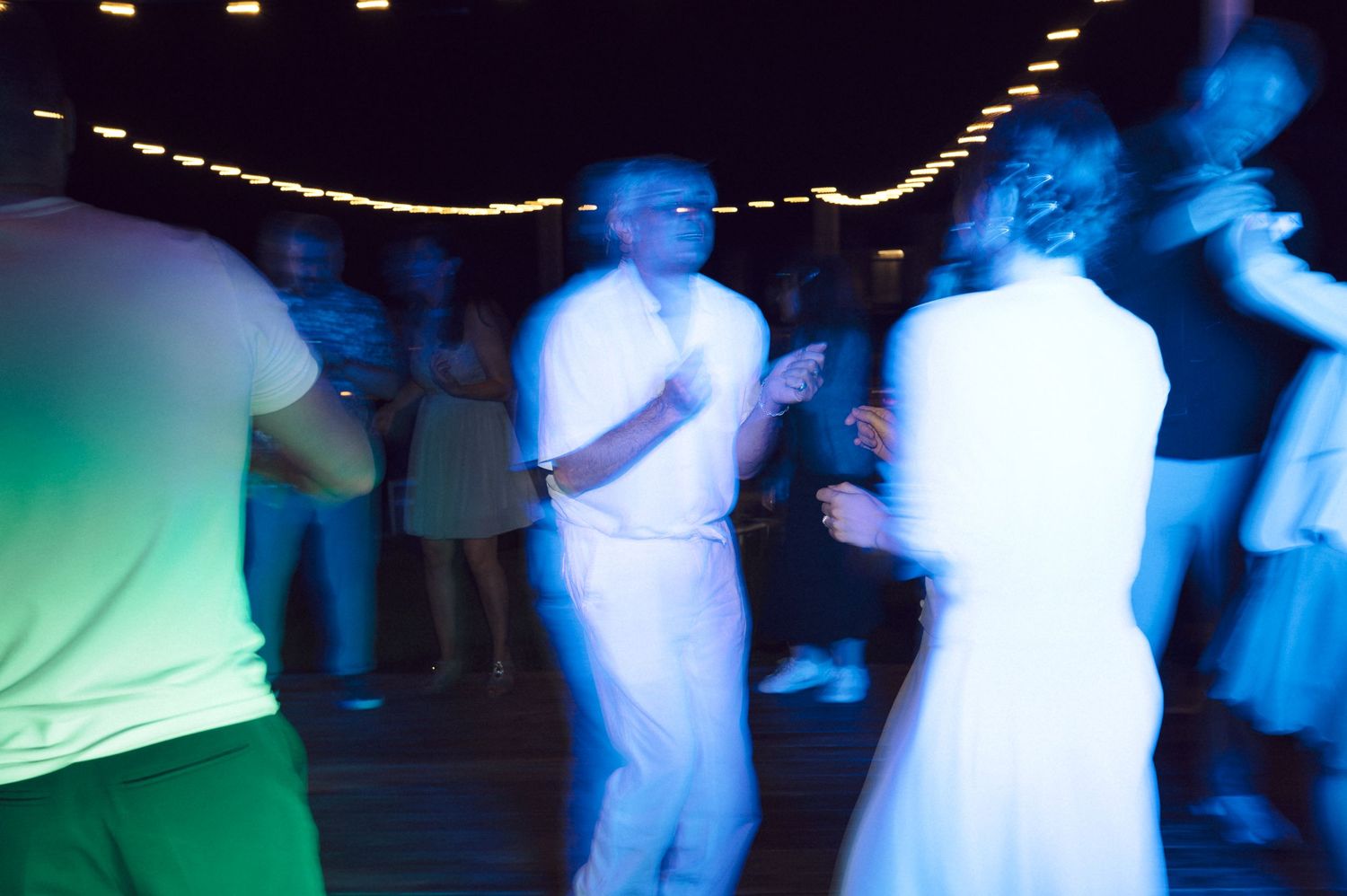 Motion-blurred scene at a nightclub or party with people dancing under blue lights and string lighting above.