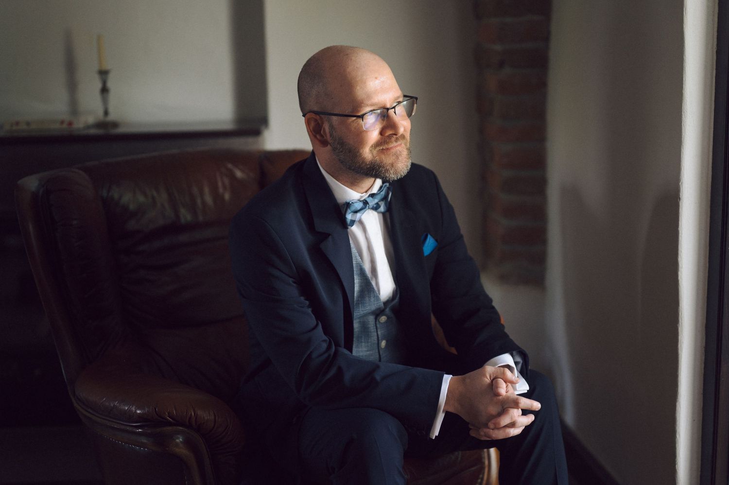 A person in a navy suit and bow tie sits thoughtfully on a leather couch near a window in natural light.
