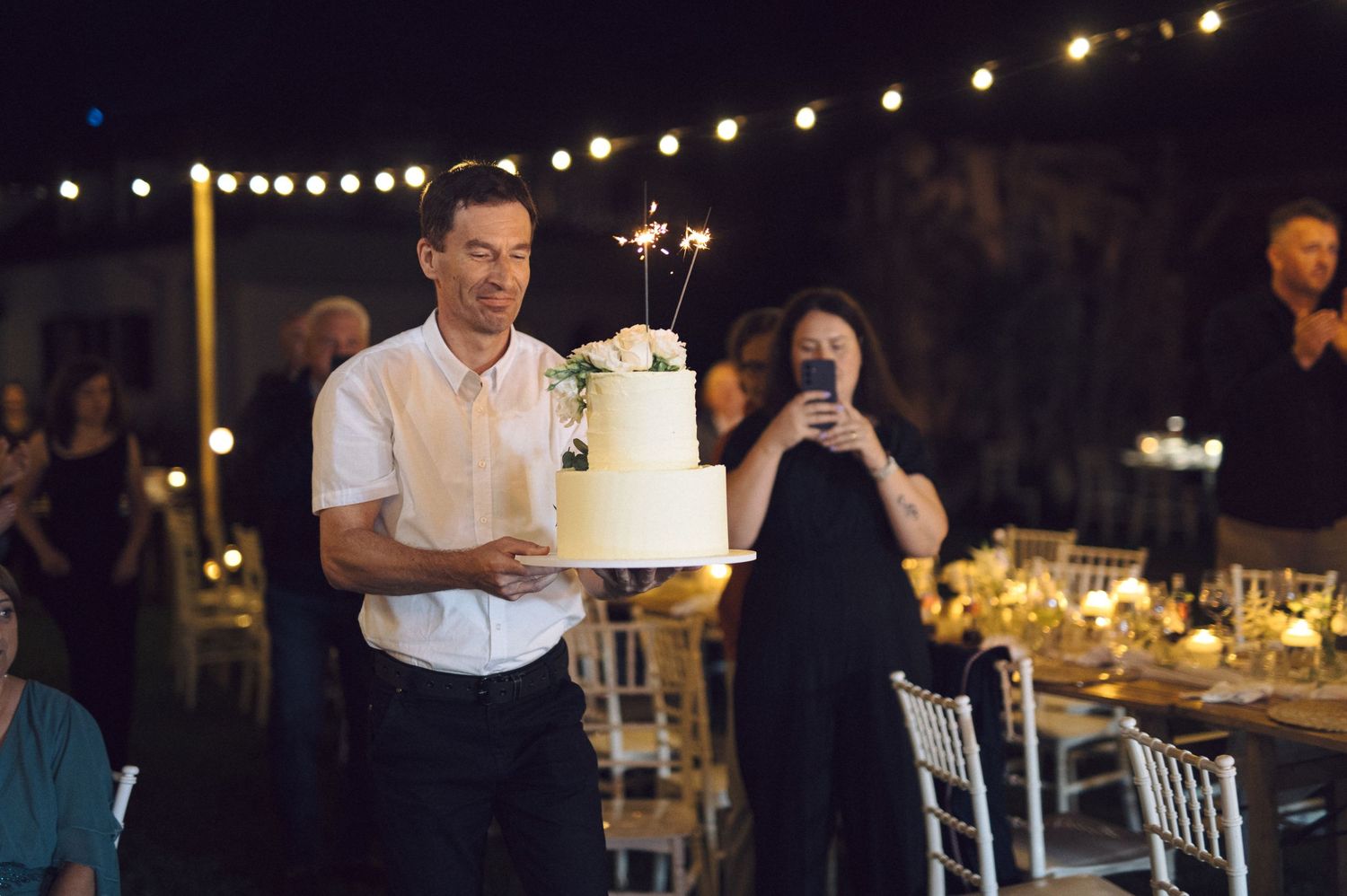 String lights illuminate an outdoor evening wedding reception with guests seated at long tables for dinner.