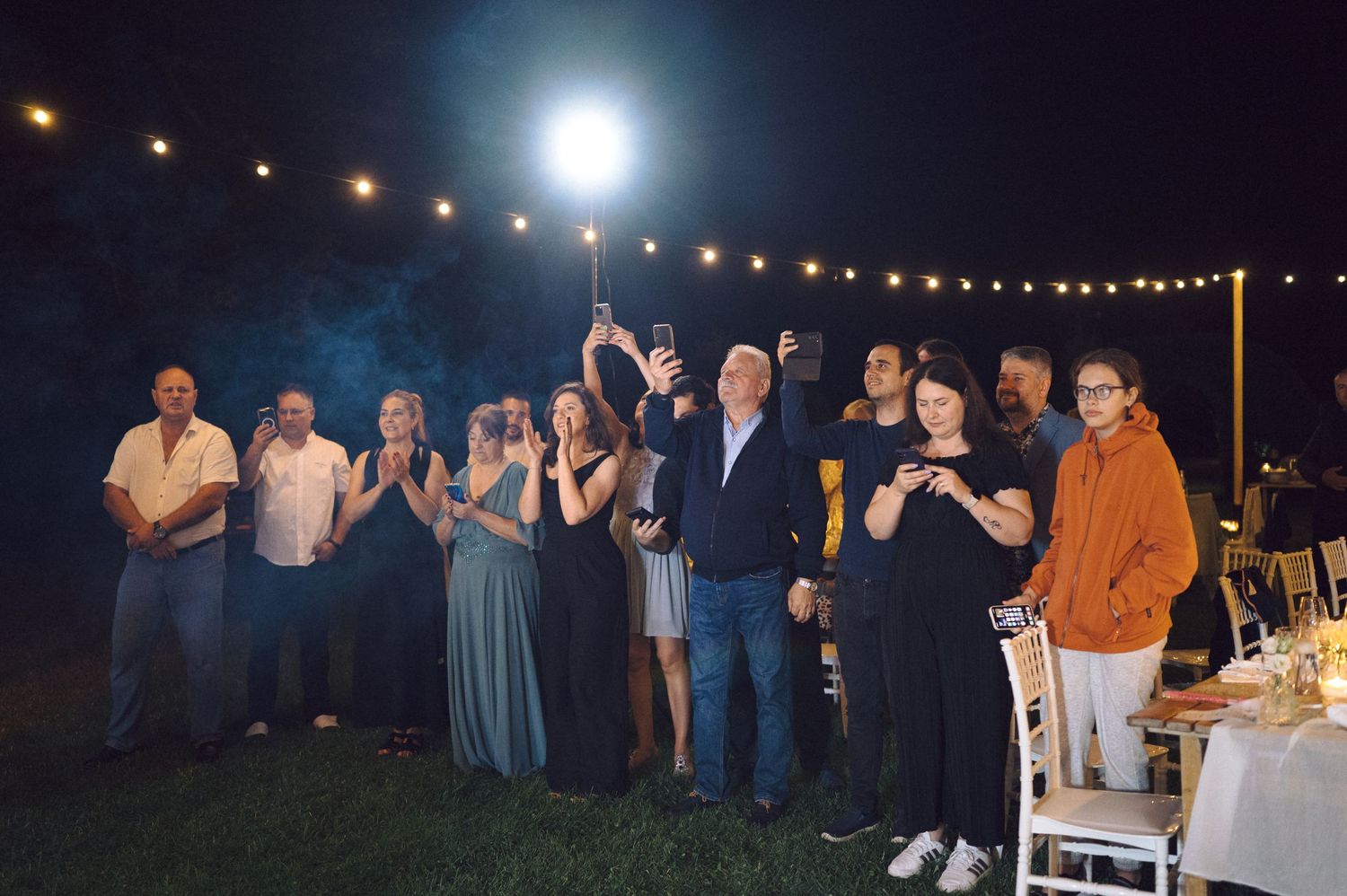 Wedding guests stand together in a row outdoors at night under string lights during an evening reception celebration.