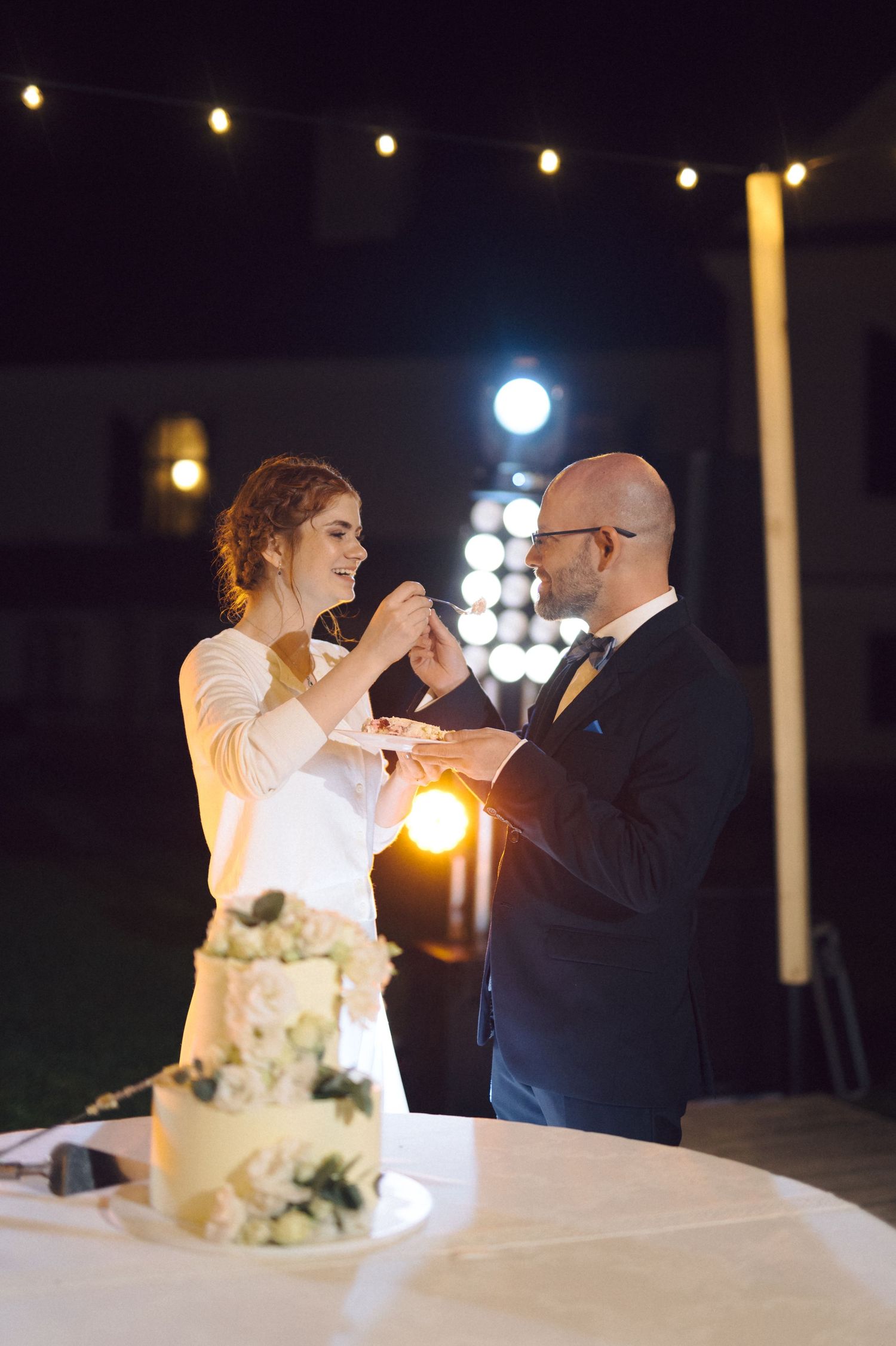 A couple stands beside their elegant white wedding cake during an intimate nighttime reception with glowing string lights.