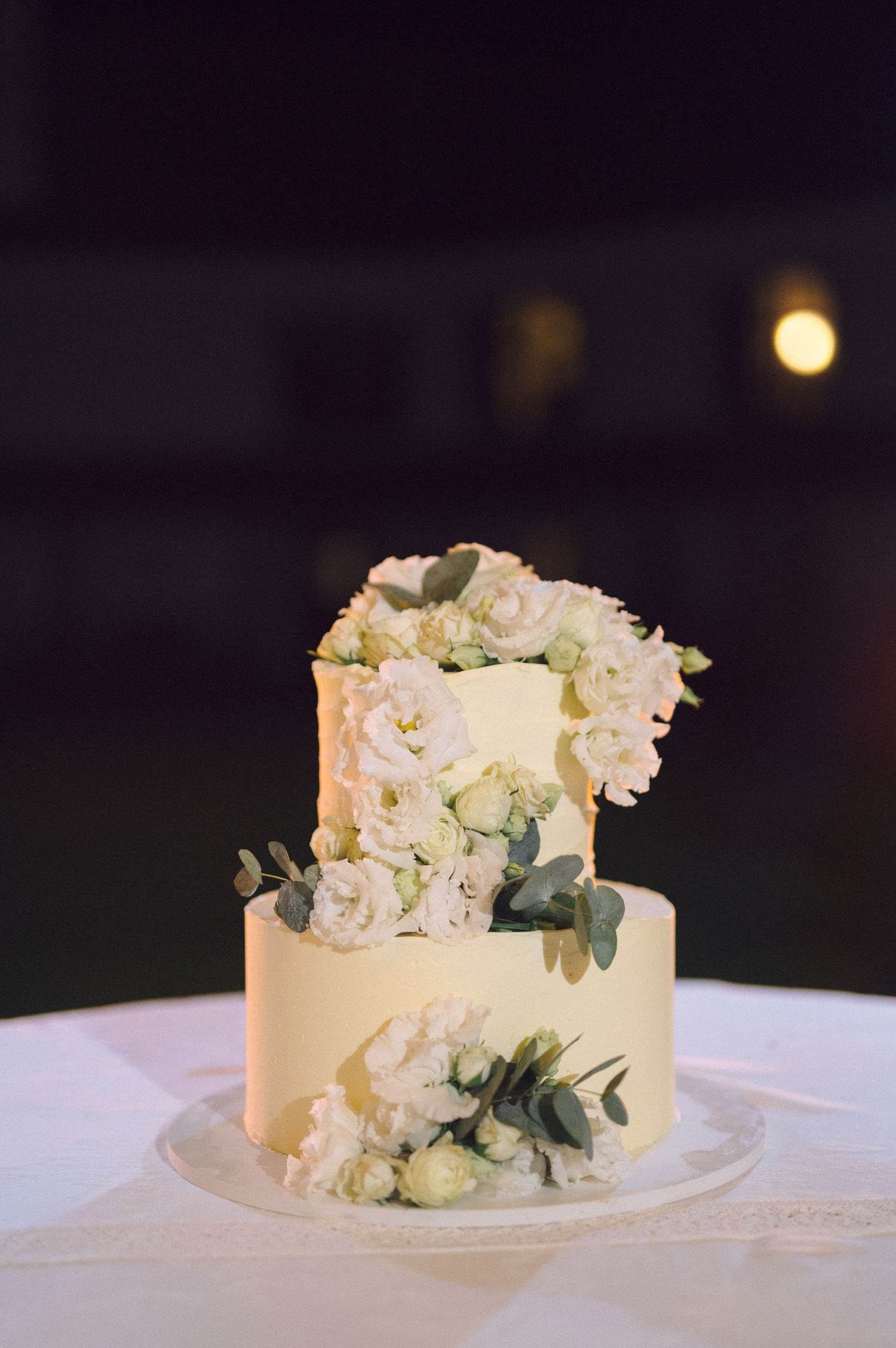 An elegant two-tier white wedding cake decorated with fresh roses and eucalyptus leaves on a white tablecloth.