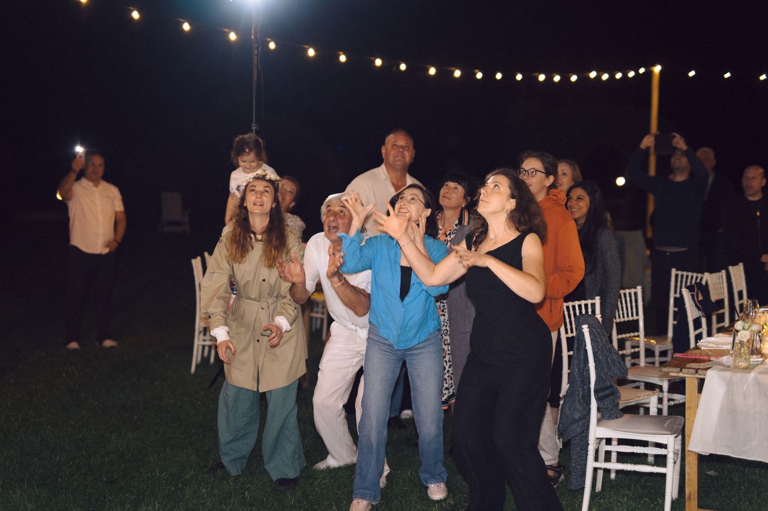 People dancing together at a nighttime outdoor event with string lights overhead and white chairs in the background.
