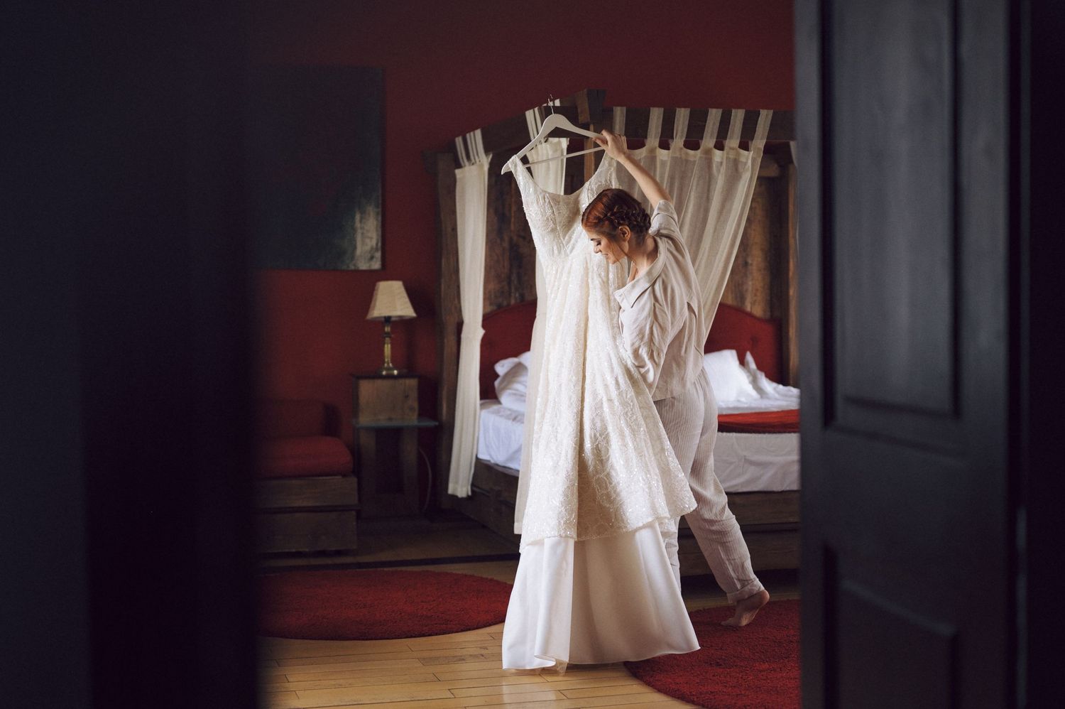 A person holds up a white wedding dress while standing in a room with red walls and elegant decor.