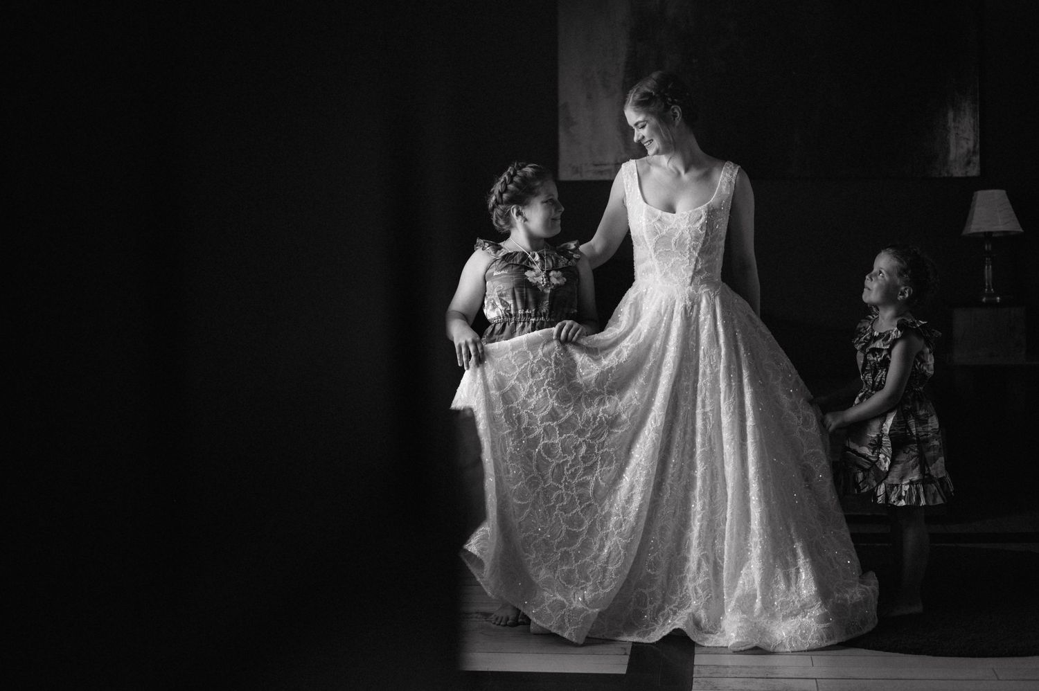 A black and white photograph shows a bride in a lace wedding dress being helped with her gown in a dimly lit room.