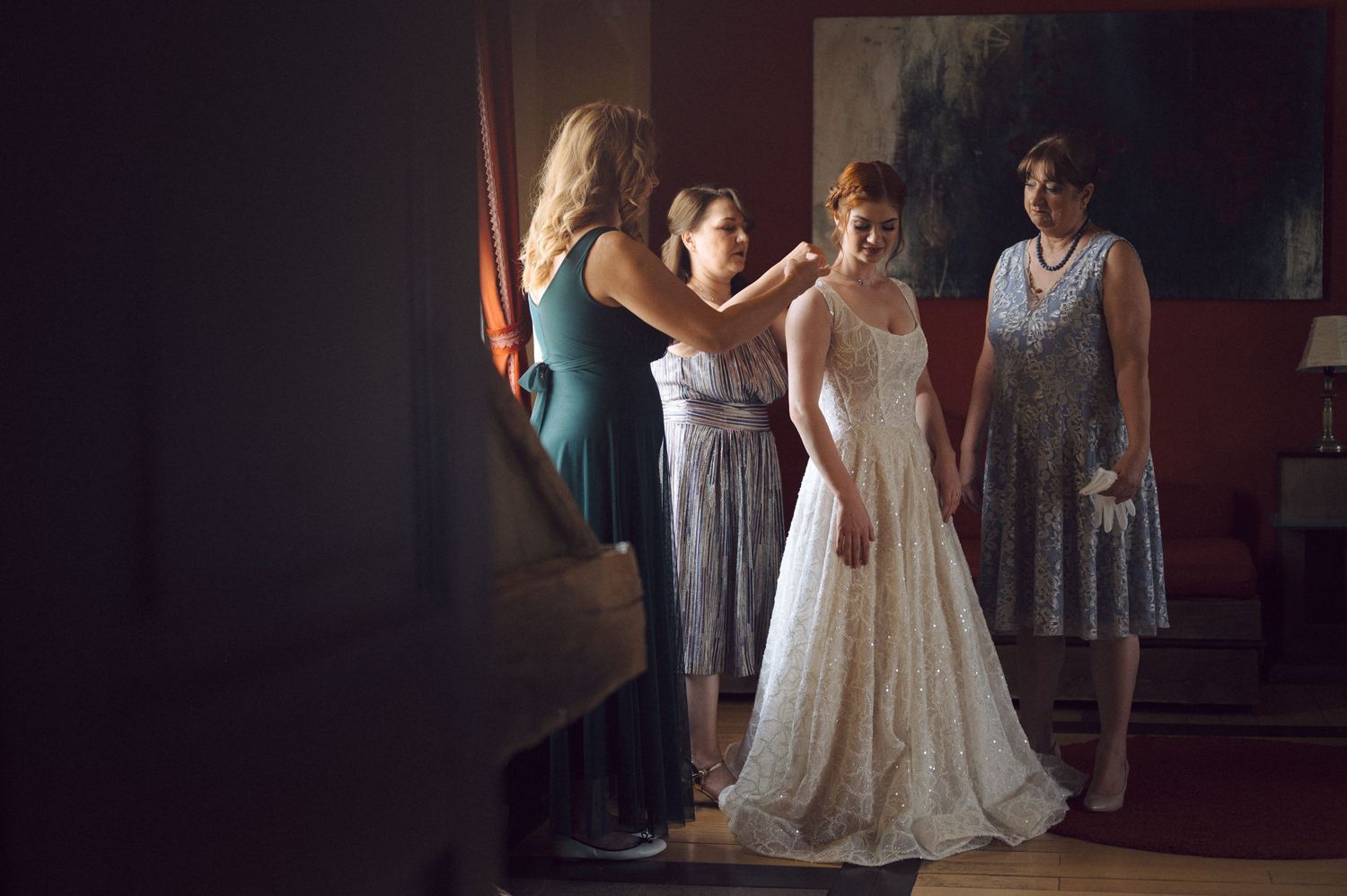A person helps adjust a white lace wedding dress while others in gray dresses look on in a dimly lit room.