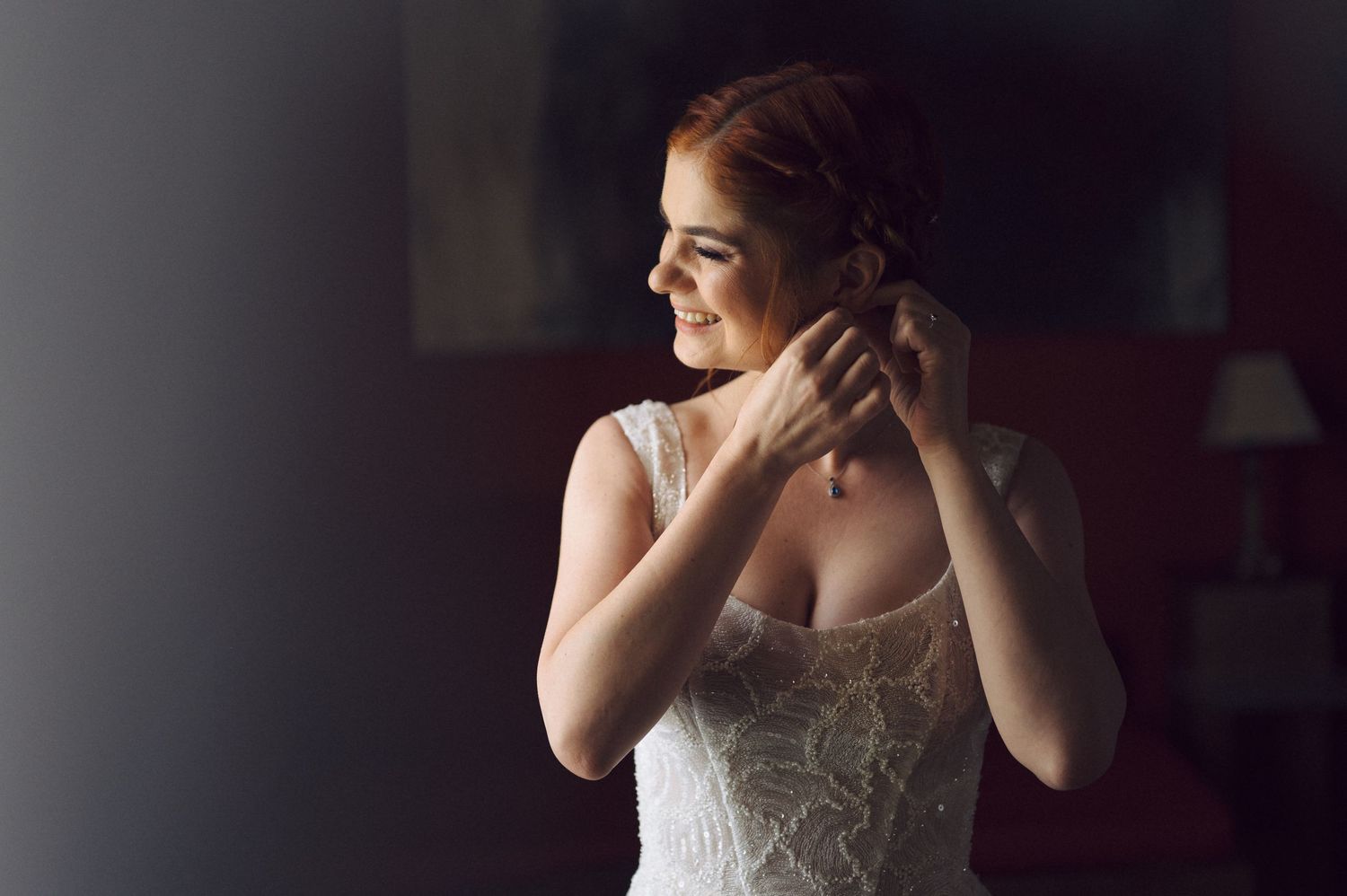 Side profile of someone in a white lace wedding dress adjusting their hair in soft natural light from a window.