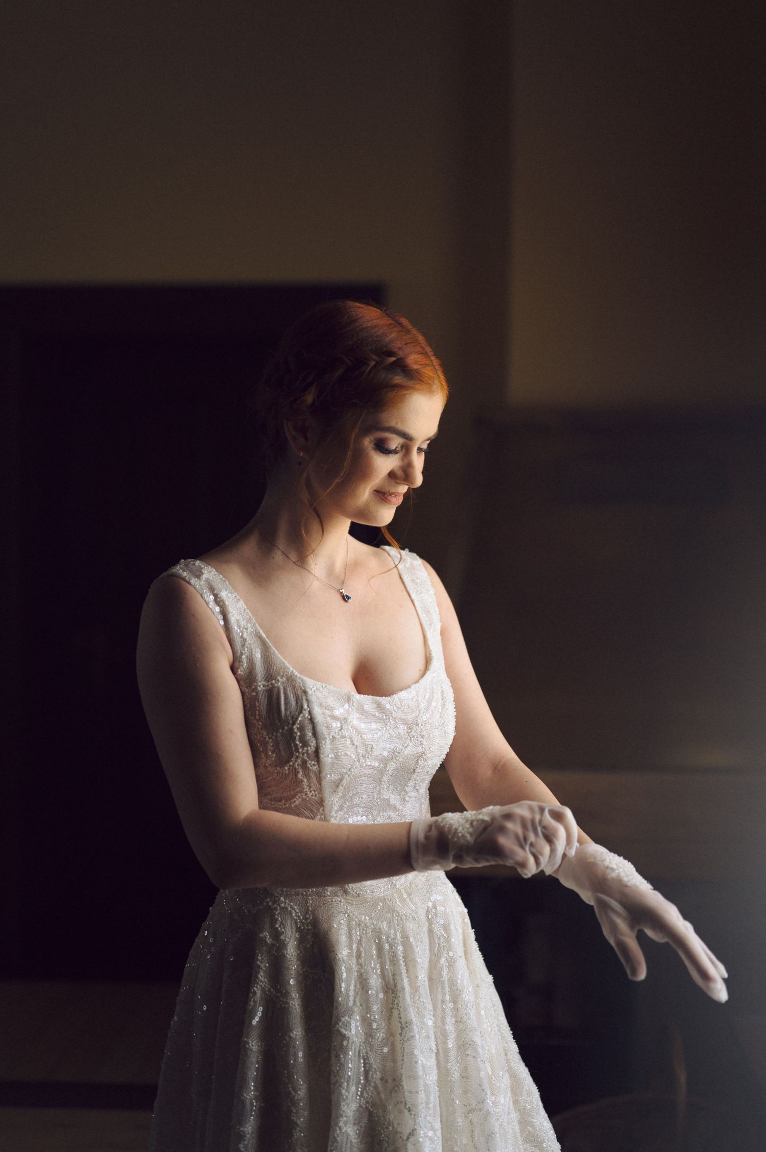 A bride in a white lace wedding dress adjusts her gloves in soft natural window light.