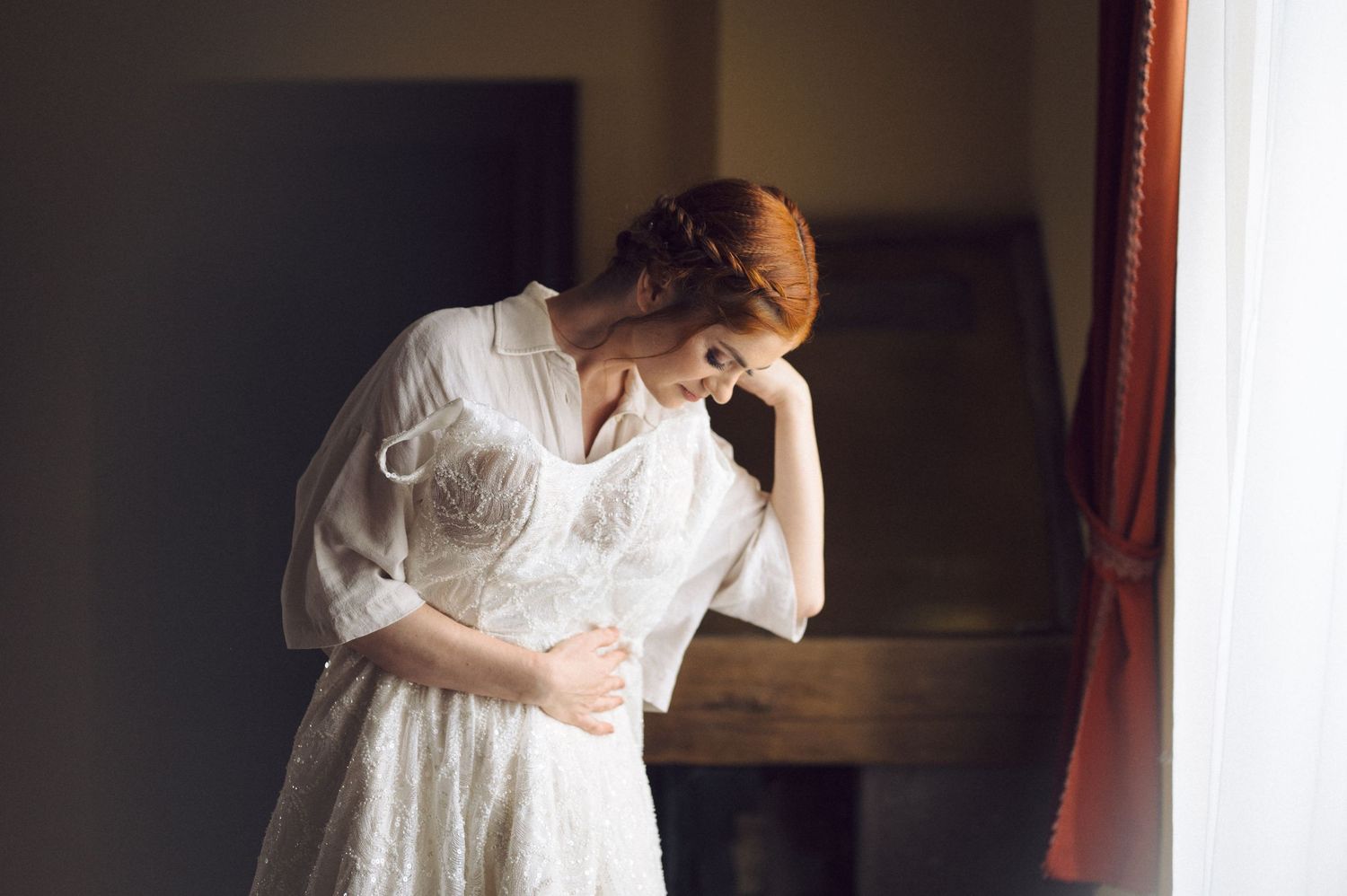 A bride in a vintage-inspired white gown and veil stands gracefully in soft window light in preparation for her wedding day.
