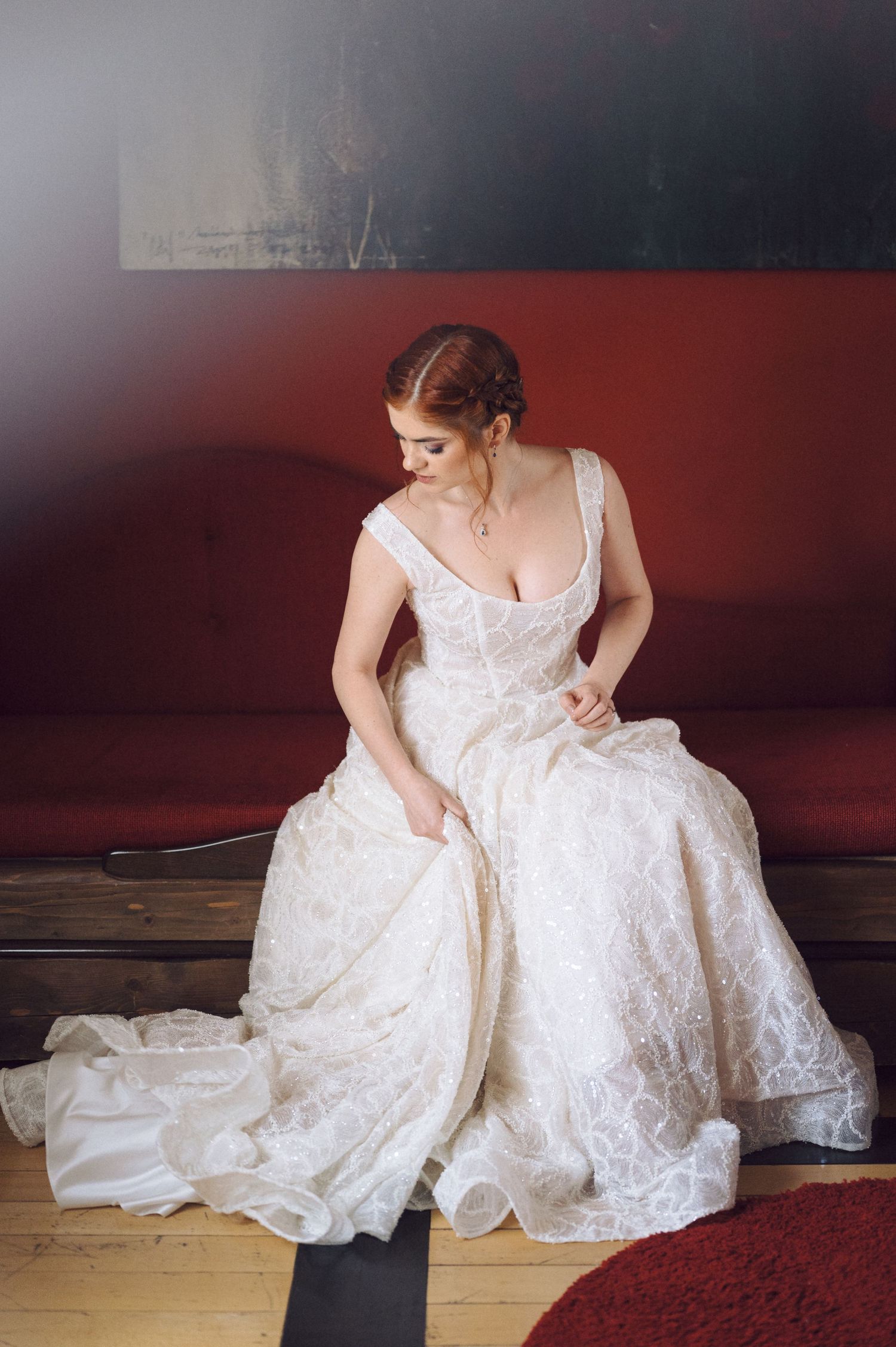 A bride in a white lace wedding dress sits gracefully on the floor against a dark red wall in an artistic photo series.
