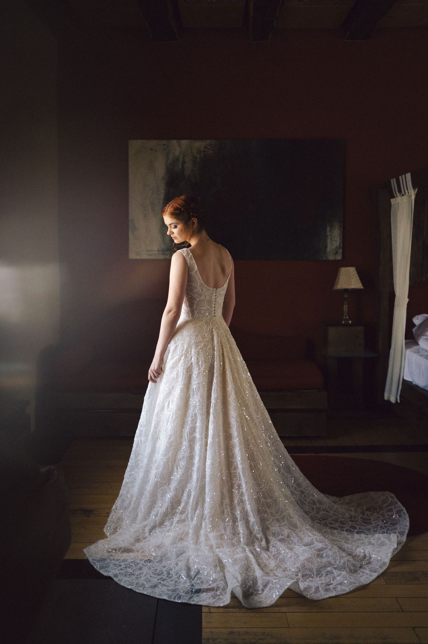 A bride in an elegant lace wedding gown with a flowing train stands gracefully in a dimly lit room.