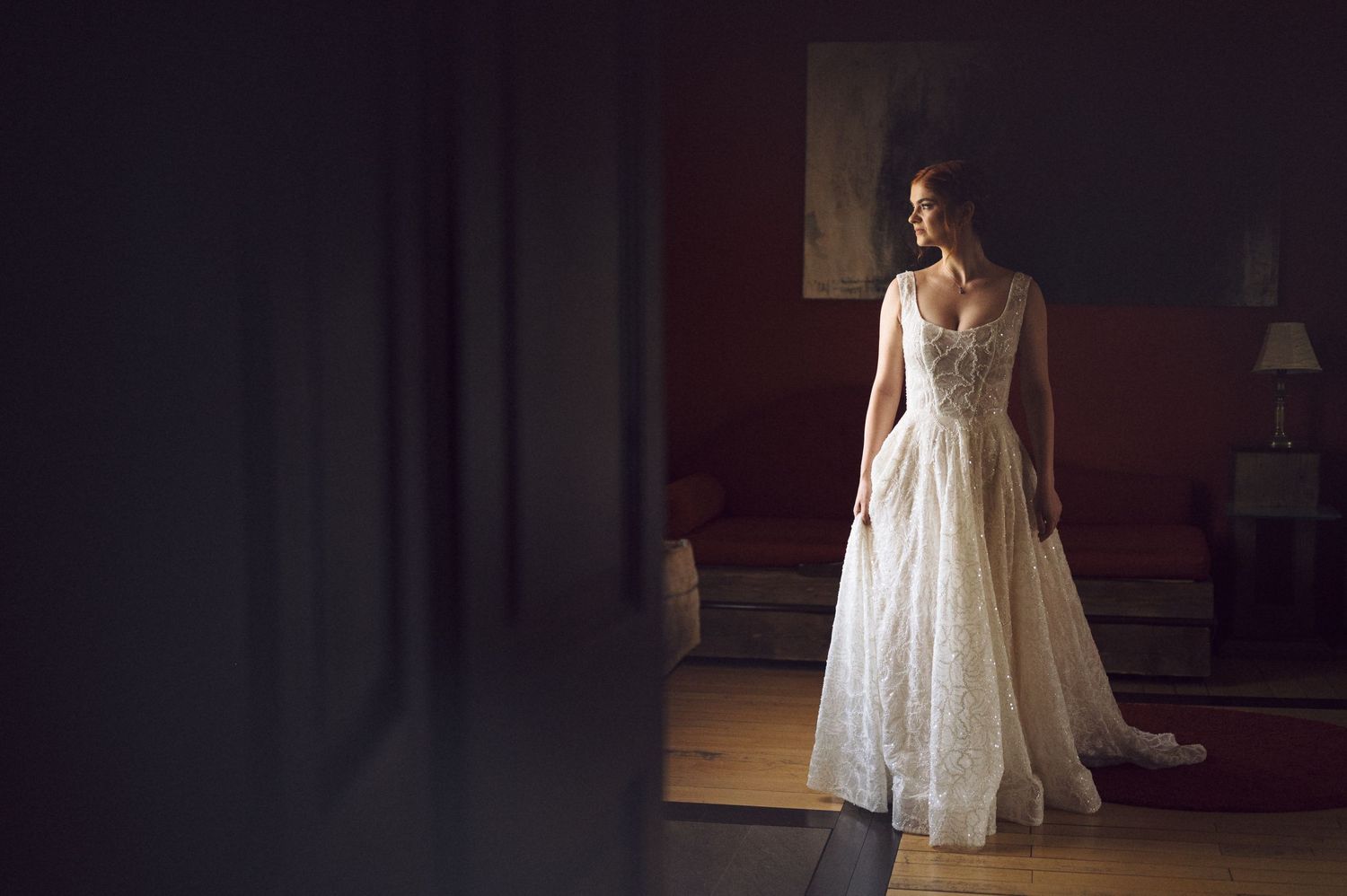 A bride stands in a dreamy white wedding dress with flowing details in a dark, moody interior setting.