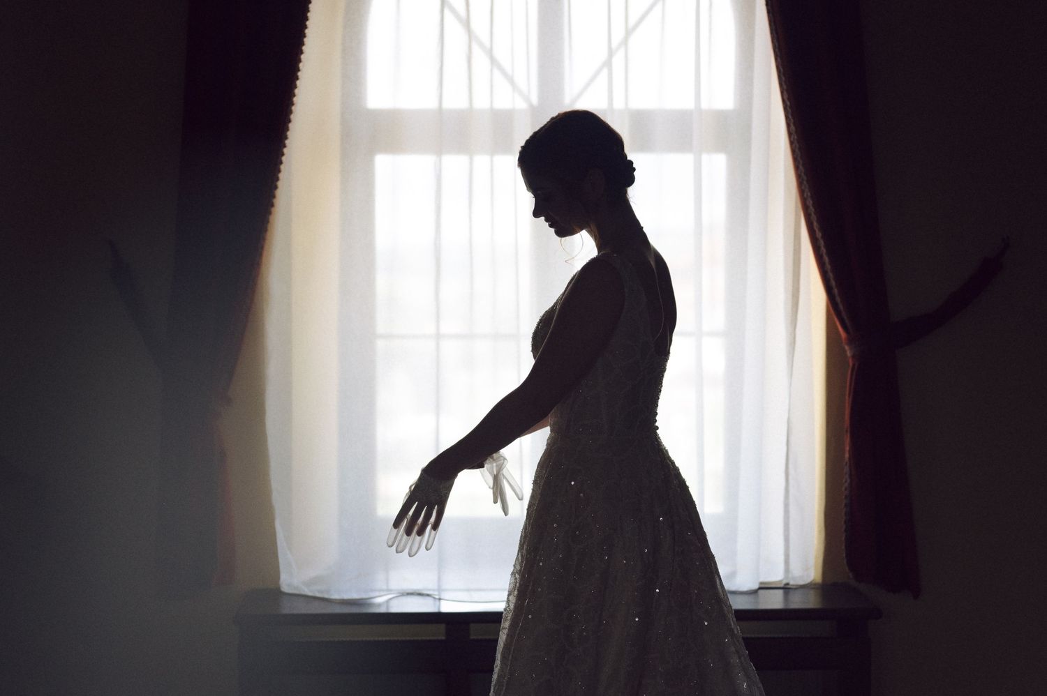 A silhouetted figure in a flowing dress stands gracefully by a bright window with sheer curtains.