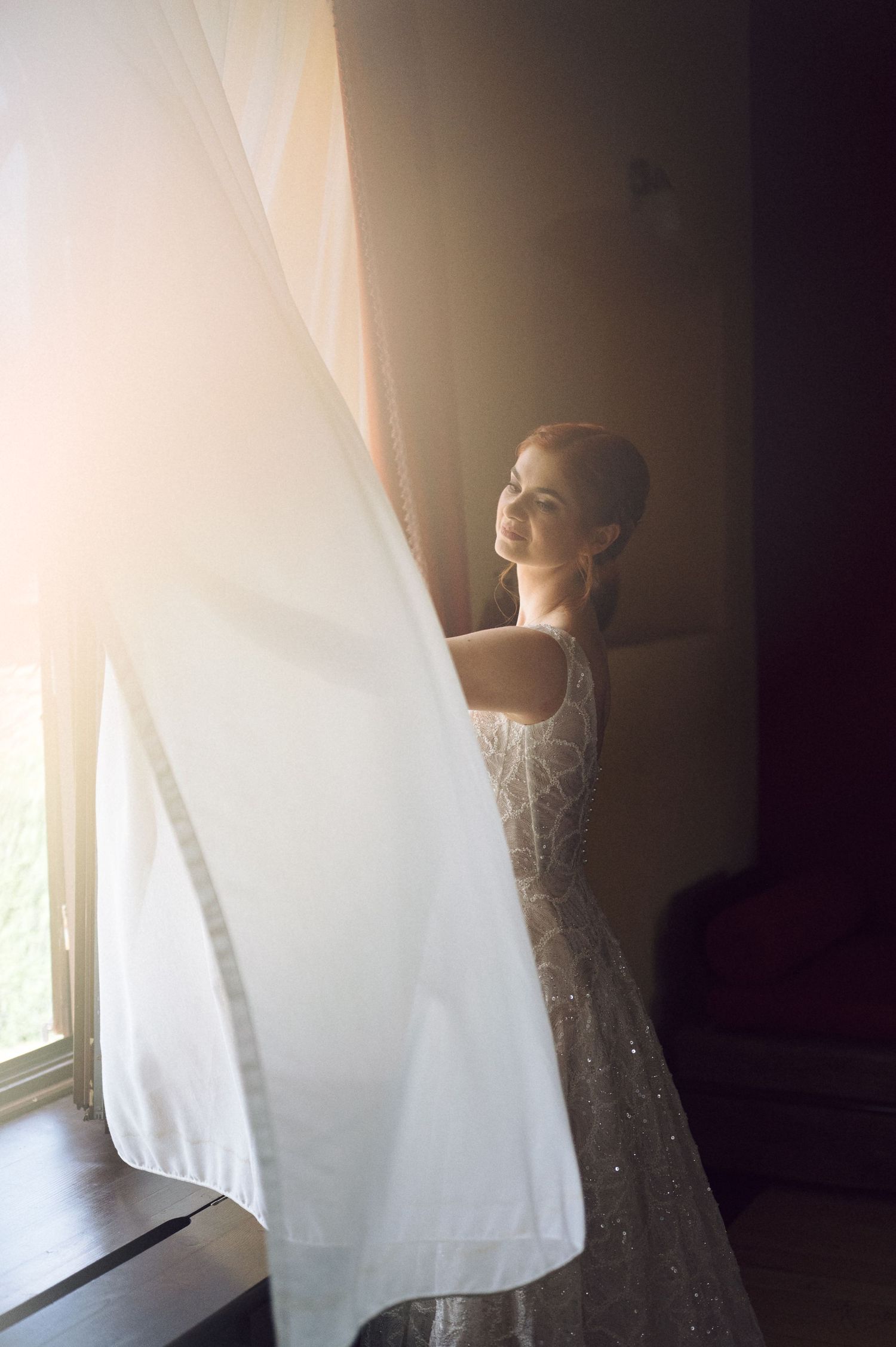 A person in a sparkly gown stands by a window holding sheer white curtains in soft natural light.