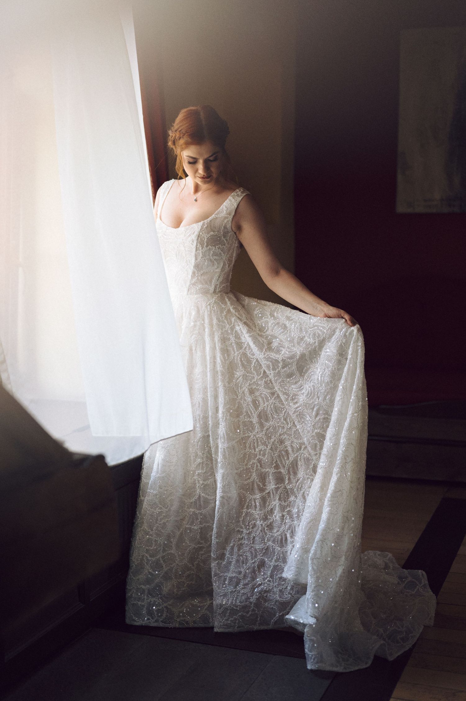 A bride in an elegant white lace wedding gown stands by a window in soft natural light.