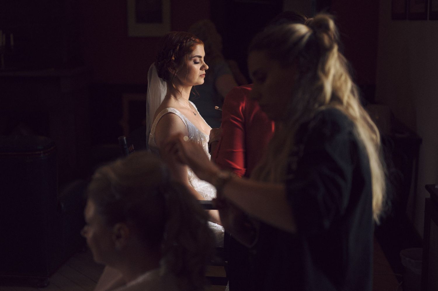 A dimly lit scene shows a figure getting ready while a stylist works in dark atmospheric lighting.