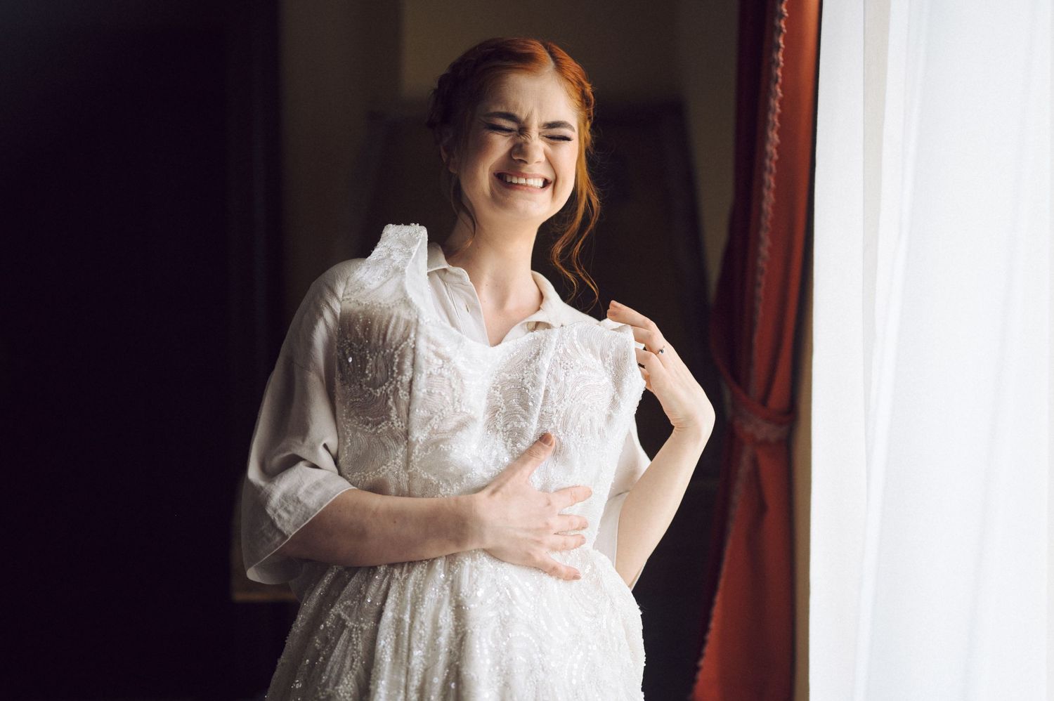 Elegant woman in white vintage lace dress stands by window with natural light creating a romantic bridal portrait.