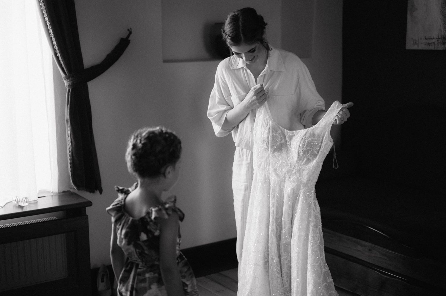 Black and white photograph showing two people looking at a white wedding dress in a minimalist interior setting.