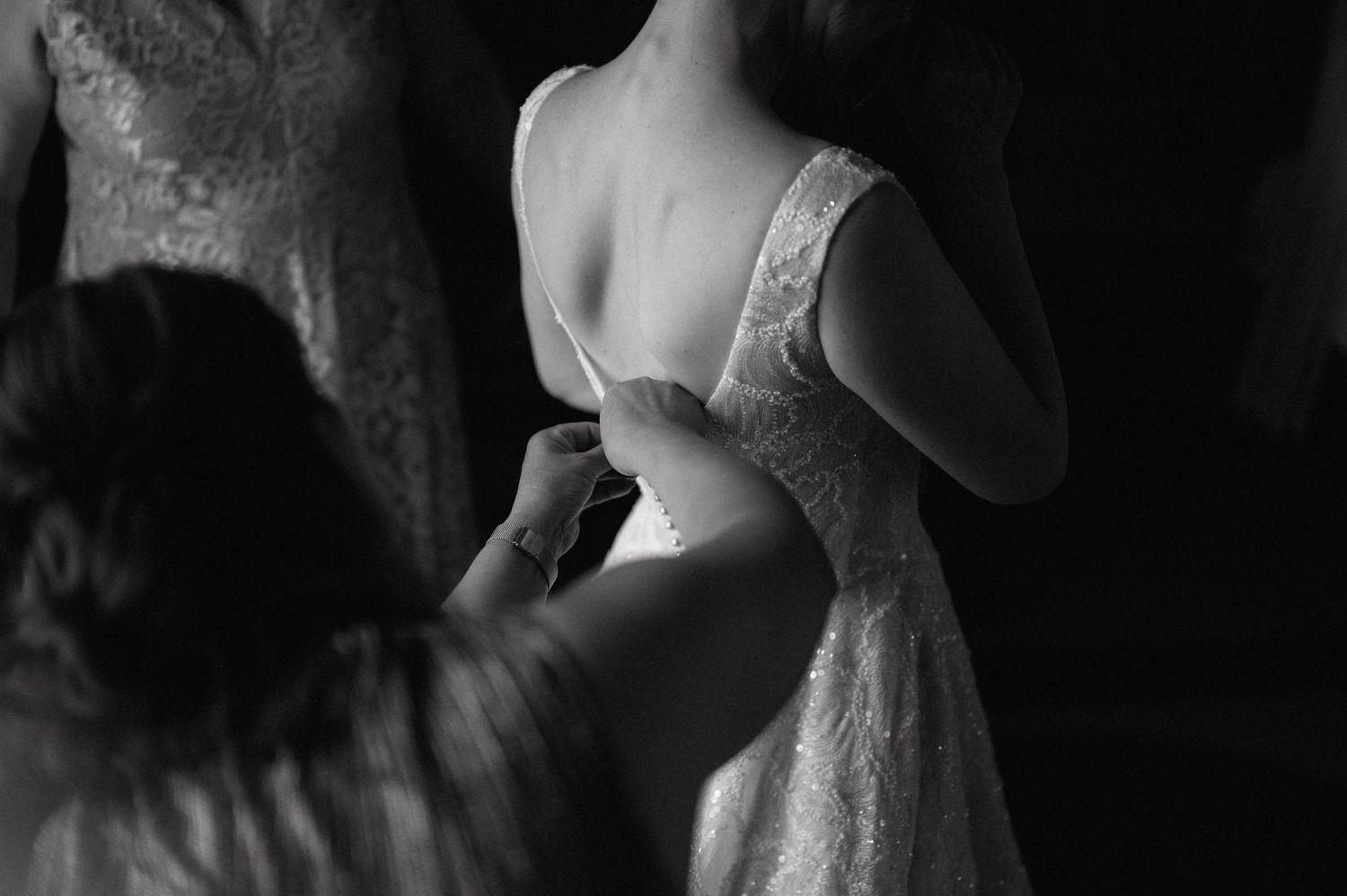 A black and white photograph shows someone being helped into a delicate lace wedding dress.