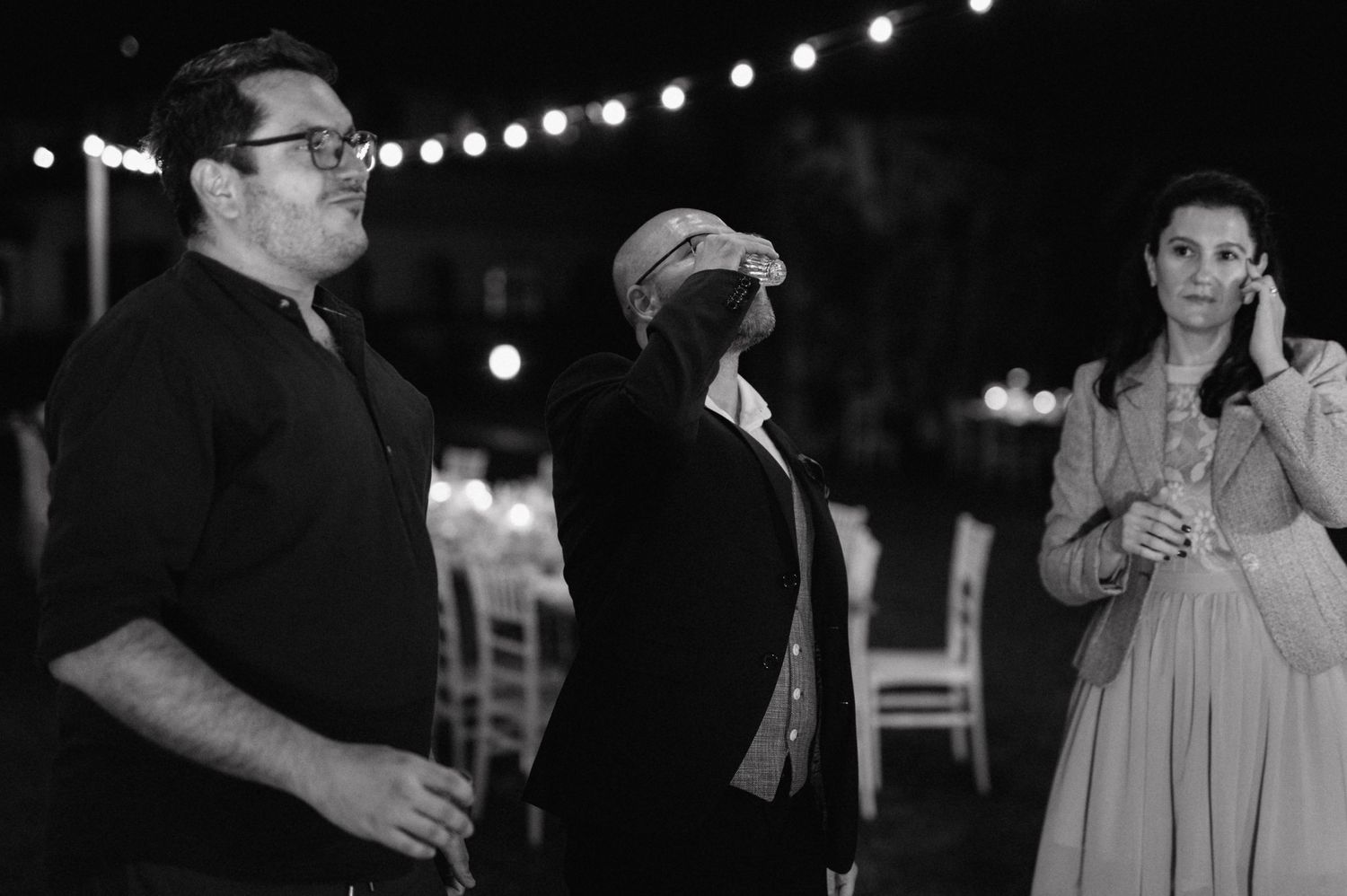 Black and white image of people chatting outdoors at night with string lights visible in the background.