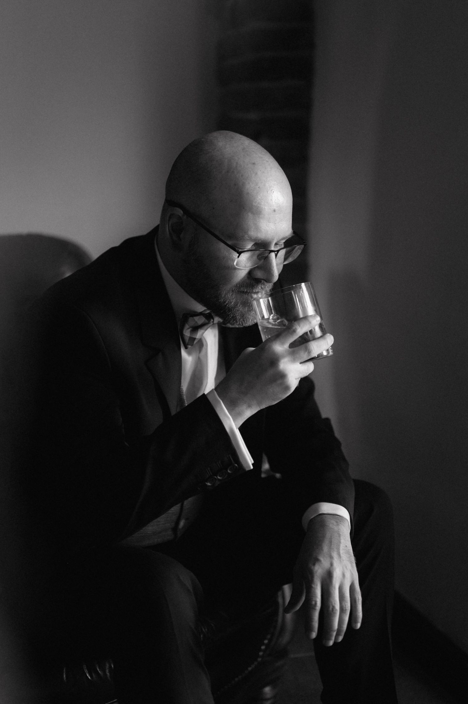 A person in a suit and glasses holds a whiskey glass in a dramatic black and white portrait photo.